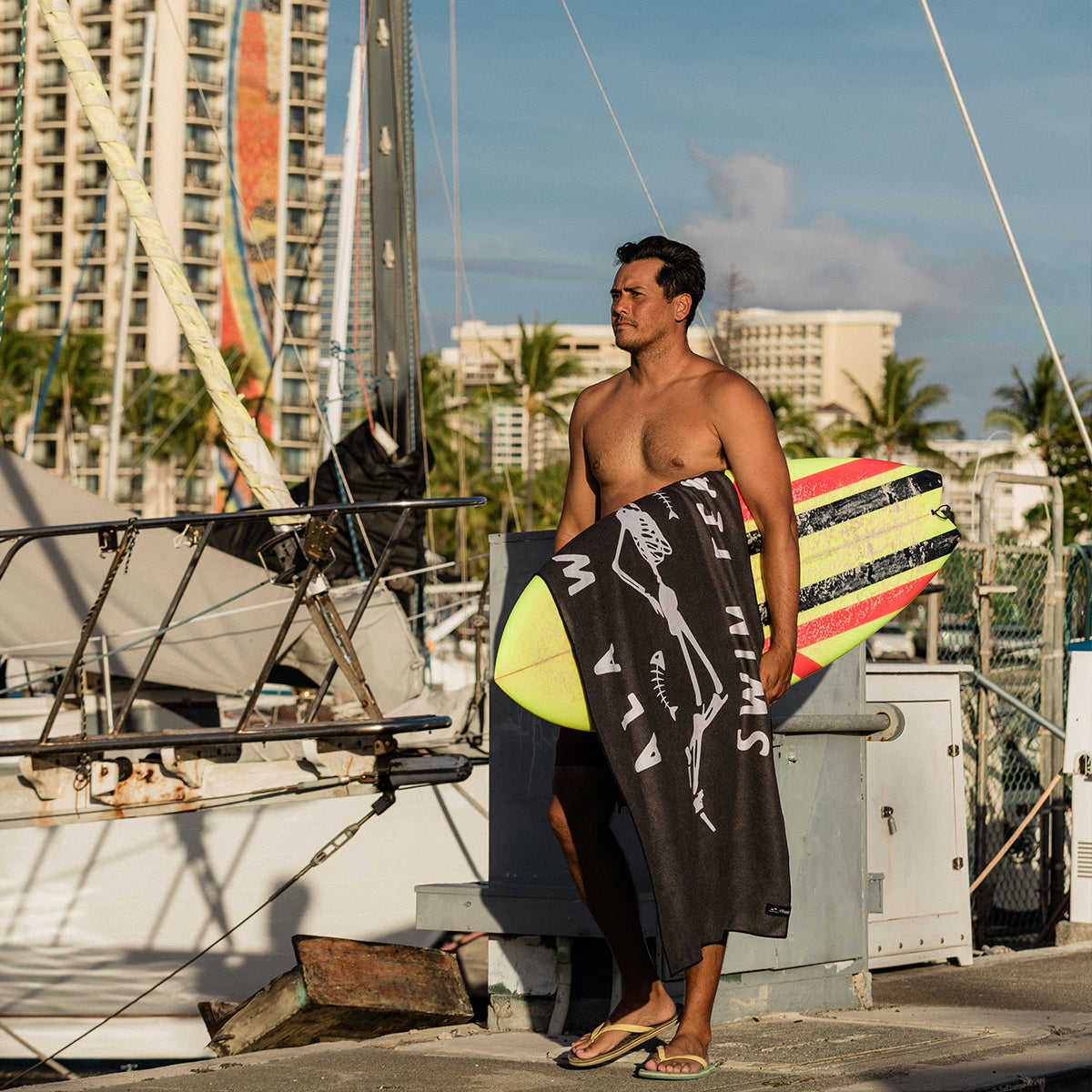 Quick-drying towel with surfboard, marina setting, sunlit day.