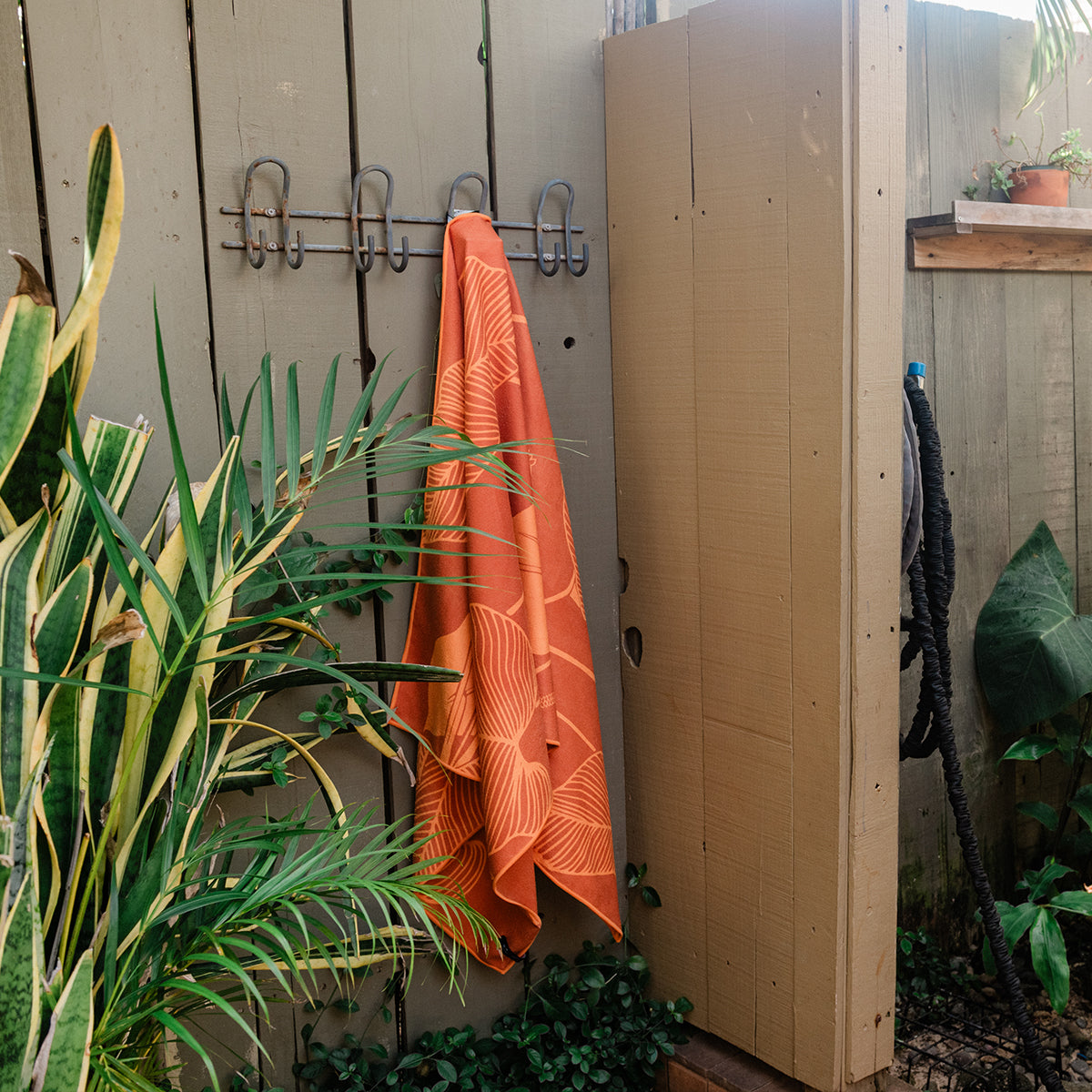 Towel with Calathea leaf pattern hanging on wooden fence outdoors near plants and shower.