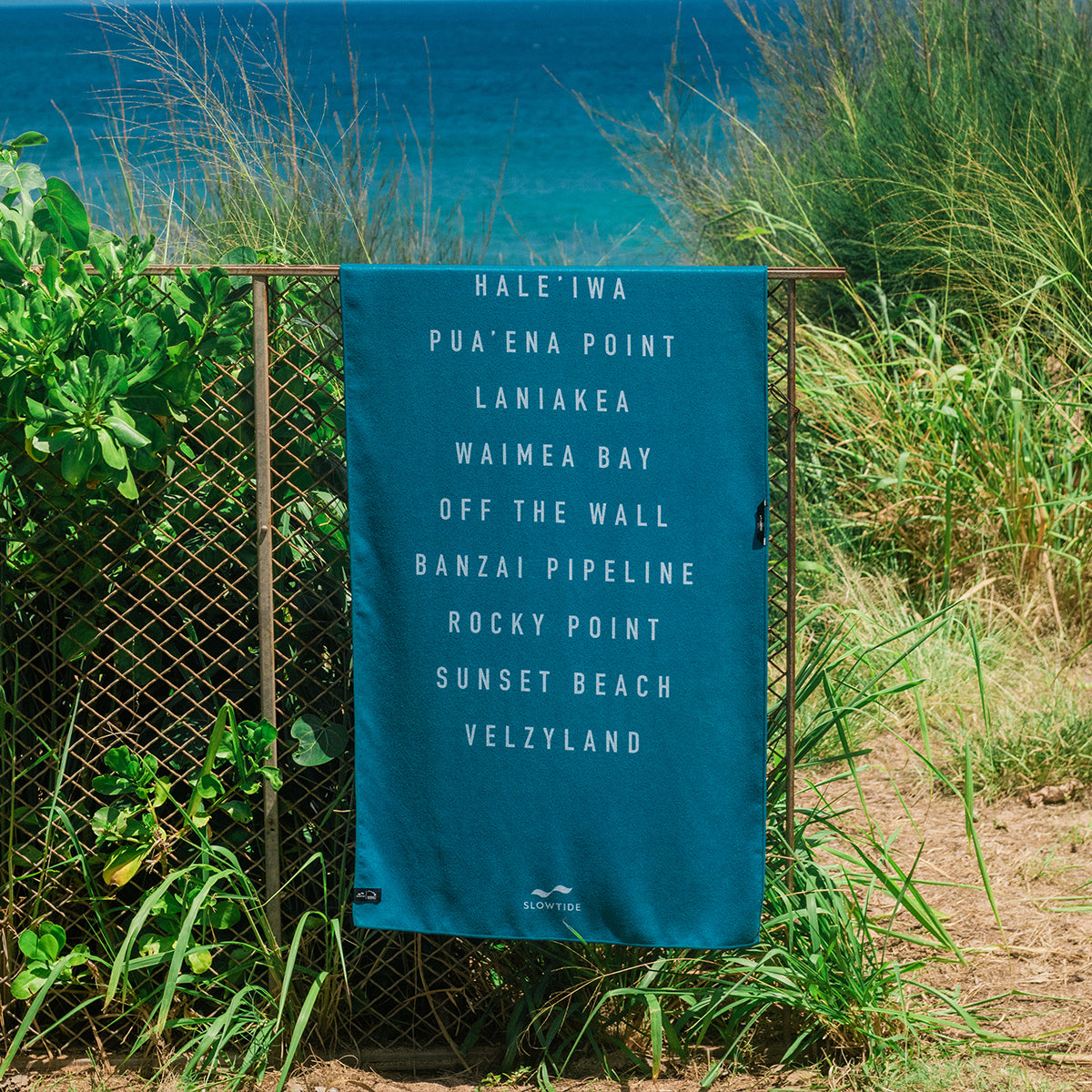 Sand-free beach towel with white Hawaiian surf spot names, displayed by the ocean.