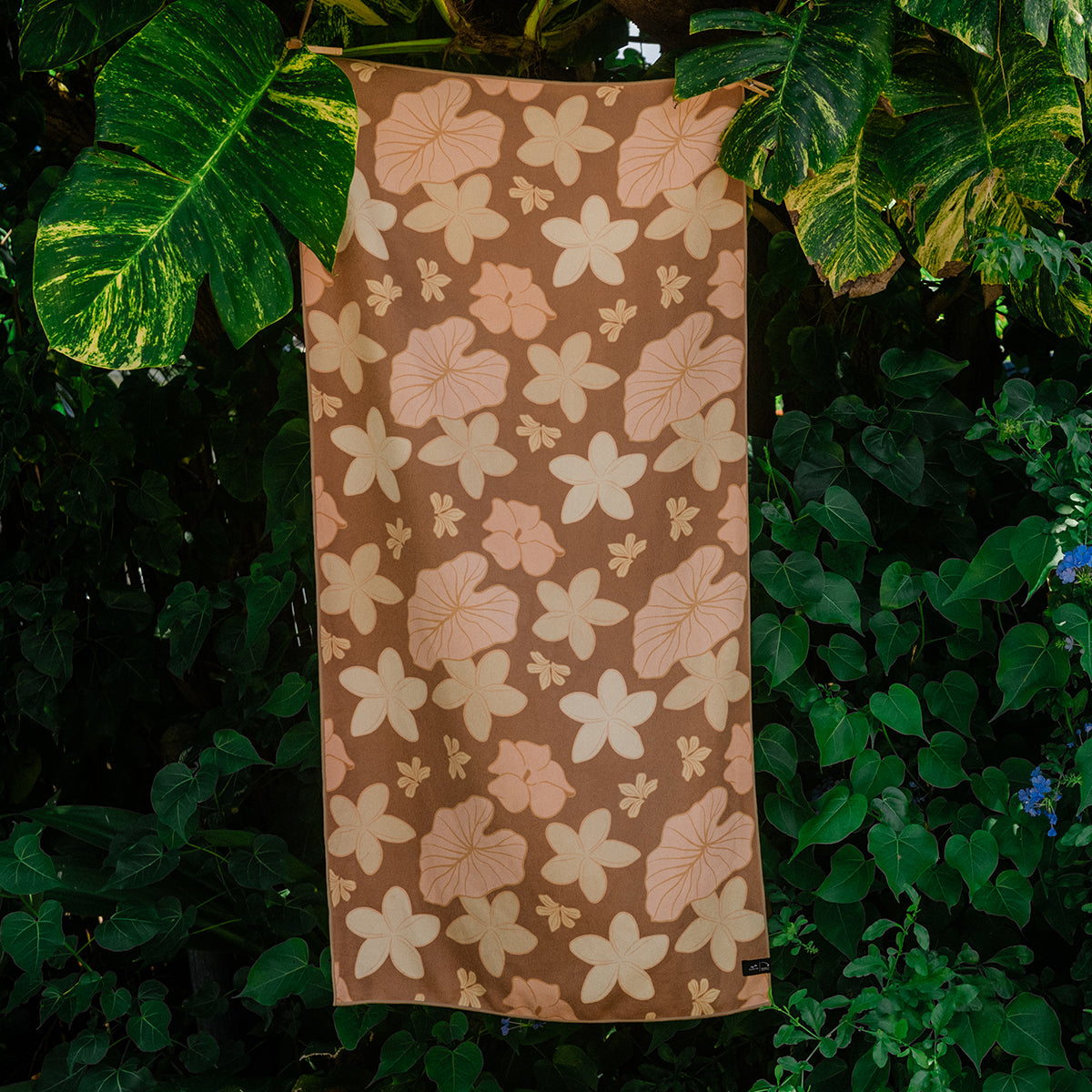 Brown sustainable towel with floral pattern hanging outside among green leafy plants.