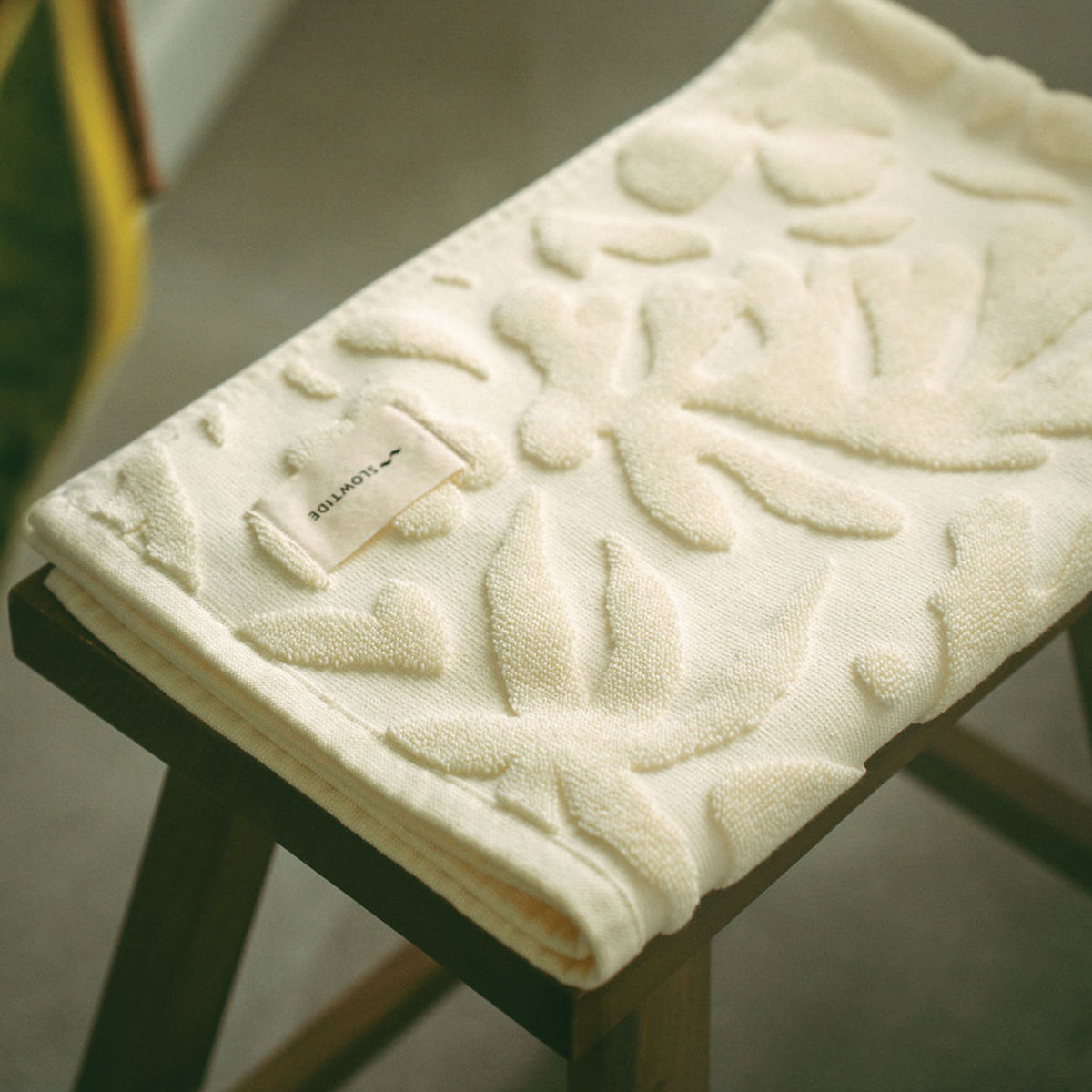 Cream cotton bath towel with raised botanical pattern on a wooden stool.