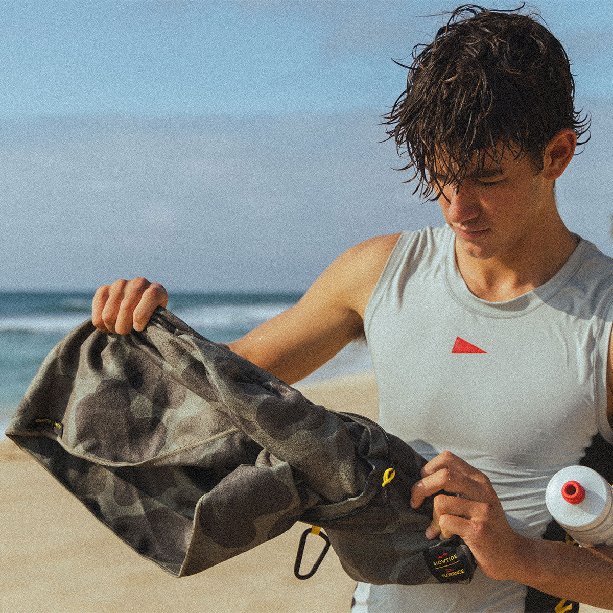 Travel towel with camo print pattern shown on a sandy beach near the ocean.
