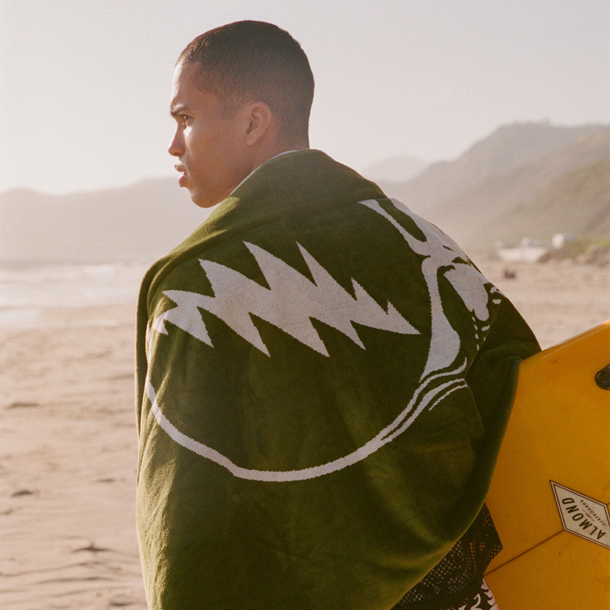 Cotton green towel with white design wrapped around surfboard on sand.