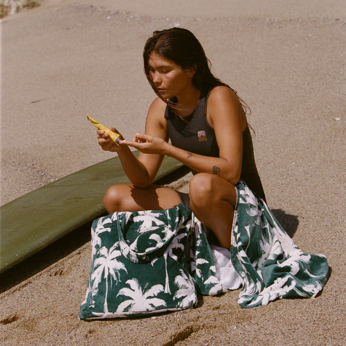 Palm print beach towel and large tote bag on sand next to surfboard.