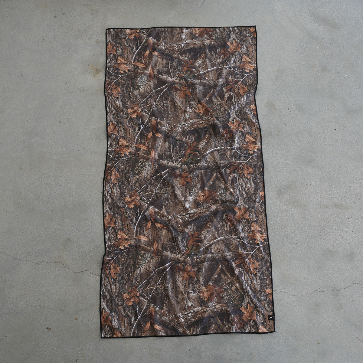 Camo quick-dry towel with brown and green leaf pattern on grey floor.