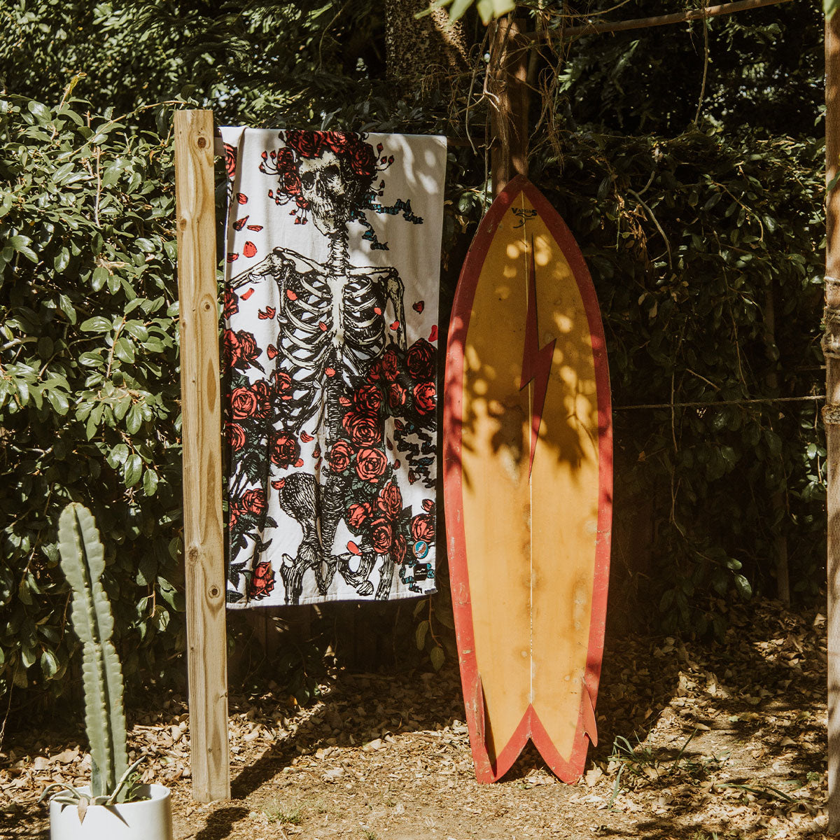 Velour beach towel with skeleton and roses print displayed beside a yellow surfboard outdoors.