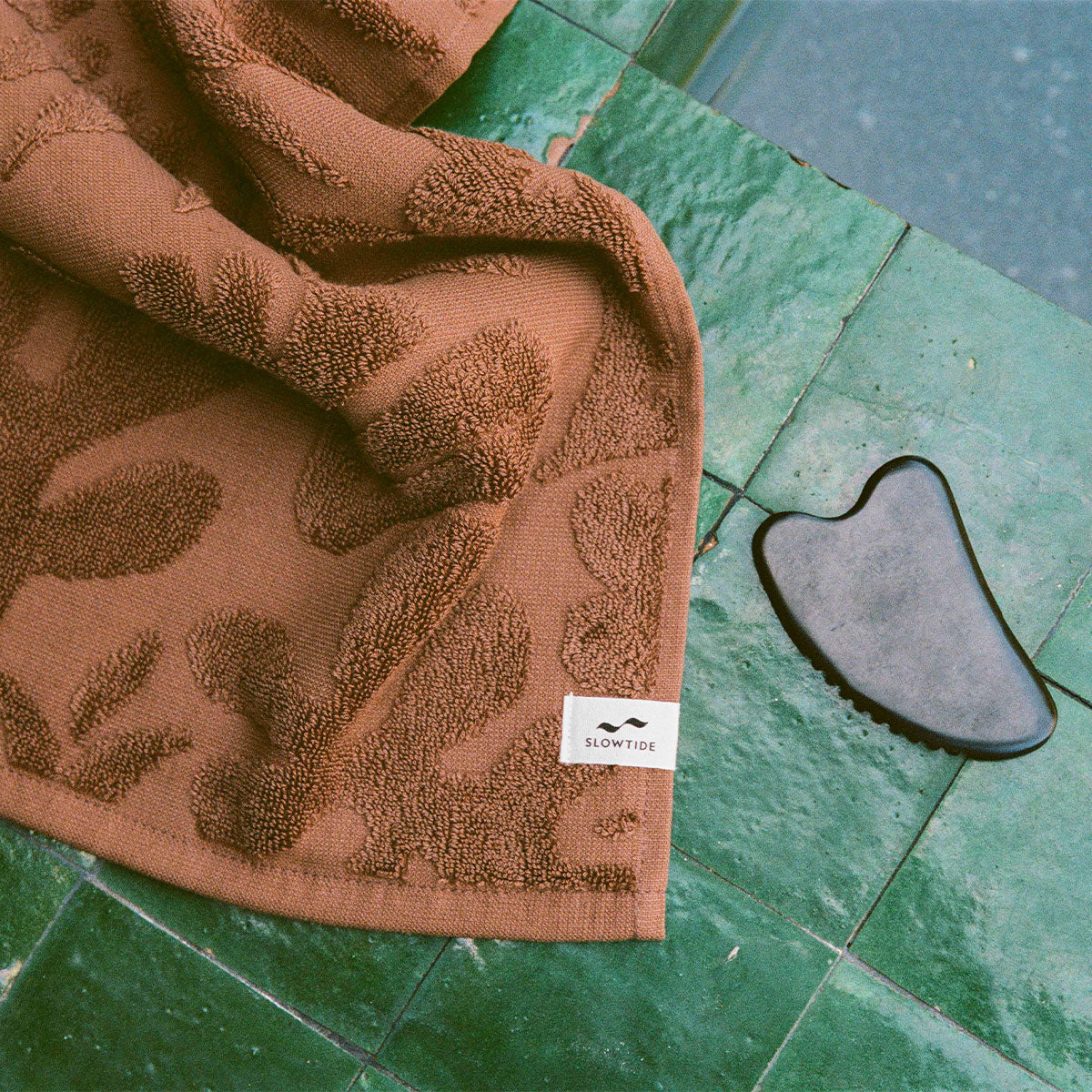 Brown 550 GSM cotton towel, absorbent, on green tiles with black gua sha stone nearby.