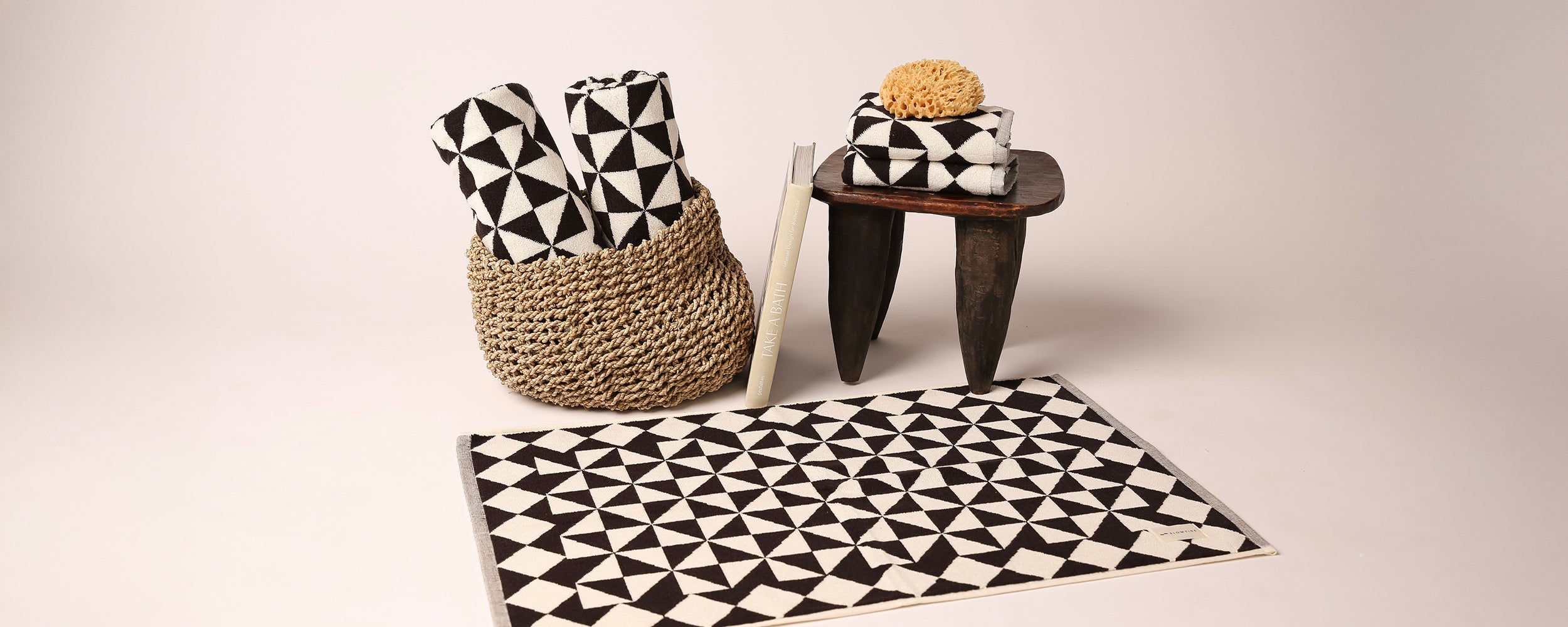 A woven basket holds rolled towels with black and white geometric patterns next to a small wooden stool topped with matching towels and a sponge; a patterned mat lies in front.