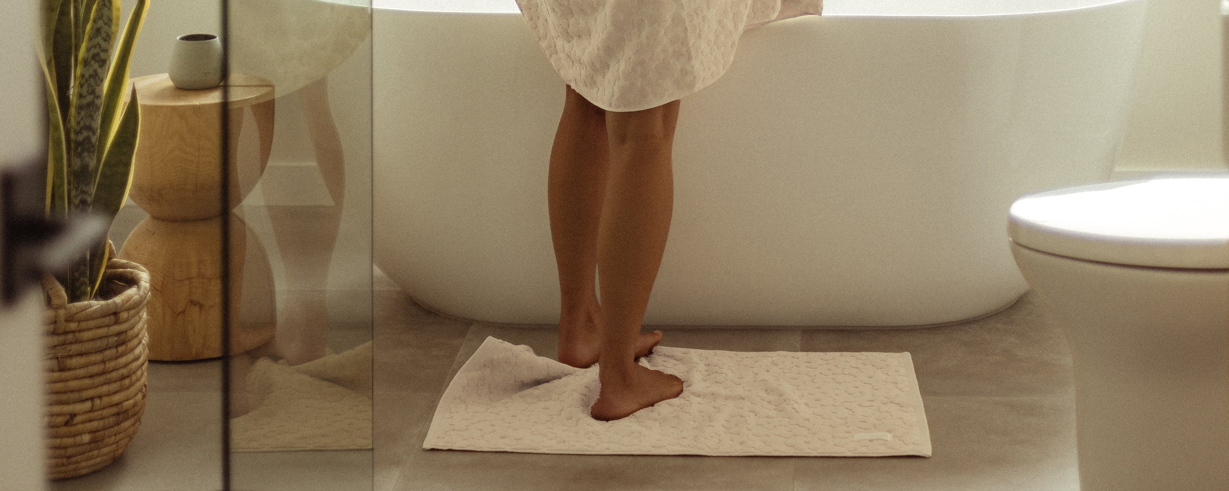 A person stands barefoot on a white bath mat in front of a bathtub, with a towel wrapped around their body in a modern bathroom with plants and natural decor.