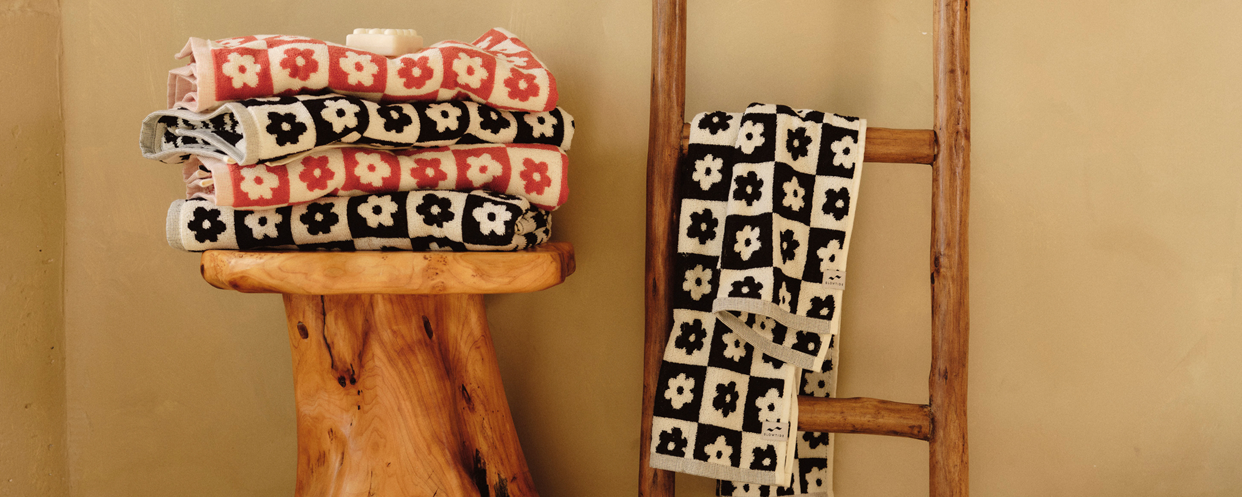 Two folded floral-patterned blankets, one in pink and cream, the other in black and cream, are displayed on a rustic wooden stool and hanging on a wooden ladder against a beige wall.