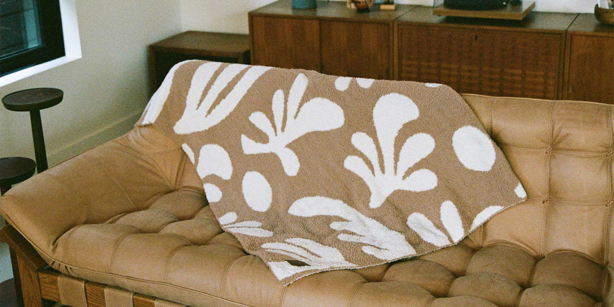 A beige tufted sofa with a tan and white abstract-patterned blanket draped over the back, in a cozy room with a wooden sideboard and modern decor.