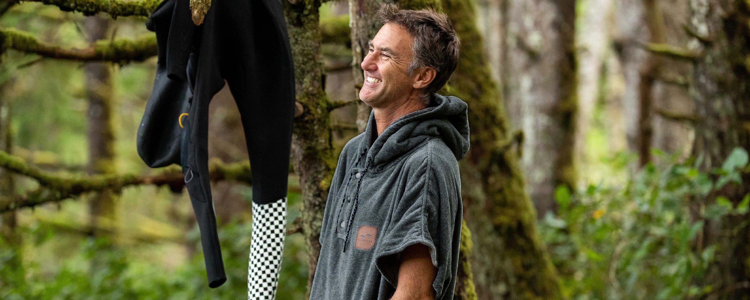 A smiling man in a hooded poncho stands in a lush, green forest with wetsuits hanging on a nearby tree branch.