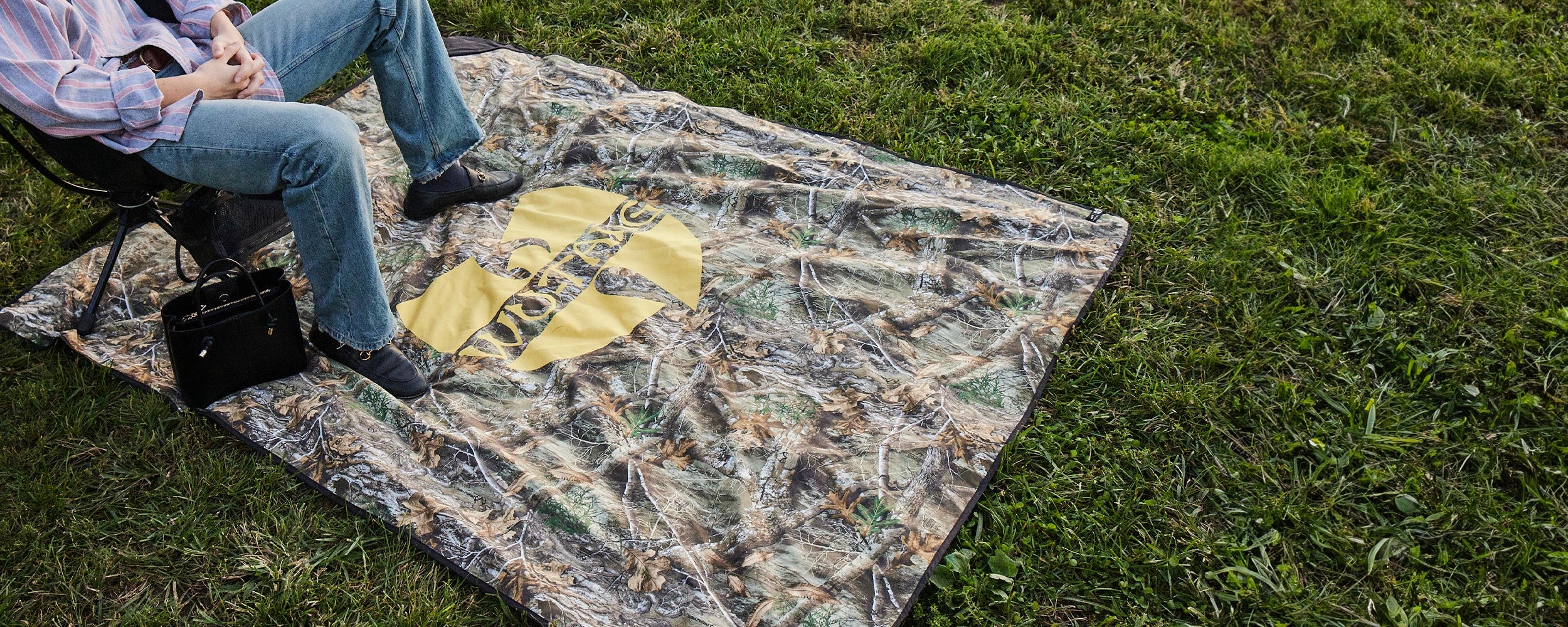 Person in jeans and boots sits on a camo-patterned blanket with a large yellow Wu-Tang Clan logo, on green grass. A black handbag is beside them.