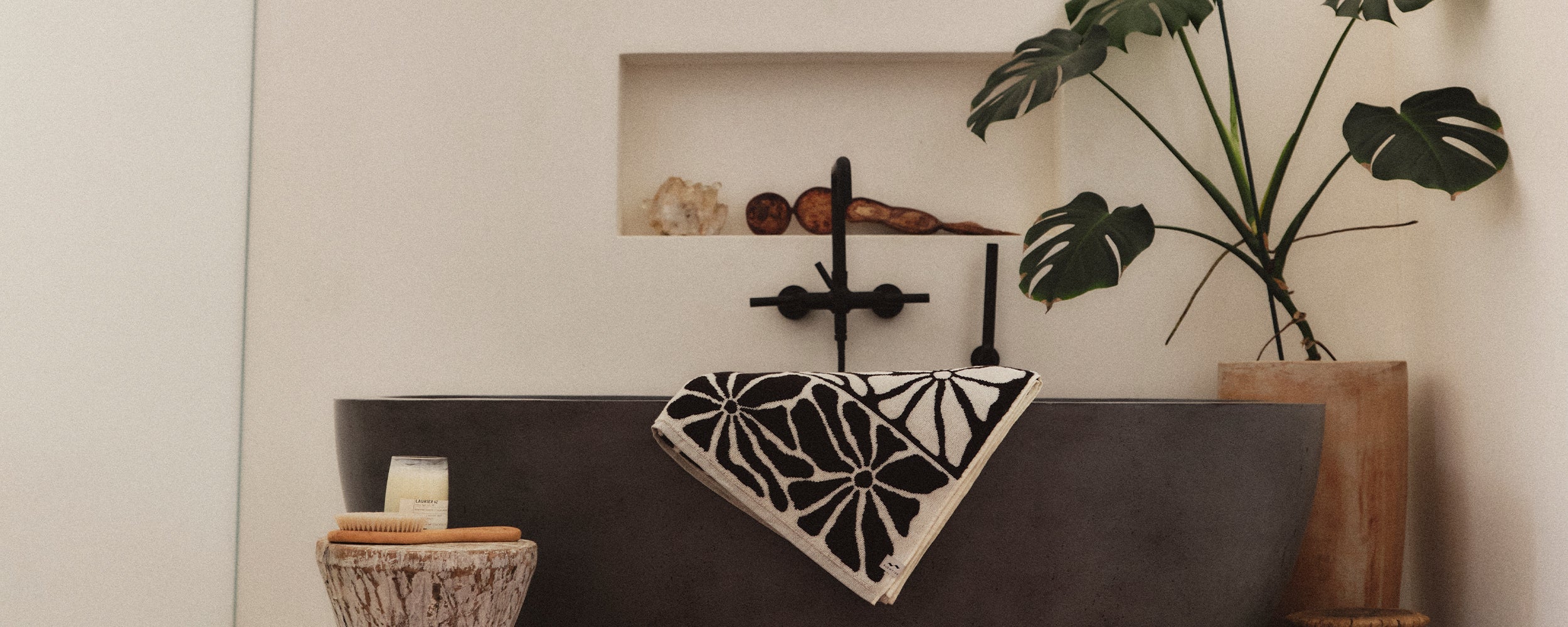Modern bathroom with a black tub, a black and white floral towel, a large potted monstera plant, a small wooden stool with a candle, and decorative shelves holding stones and shells.
