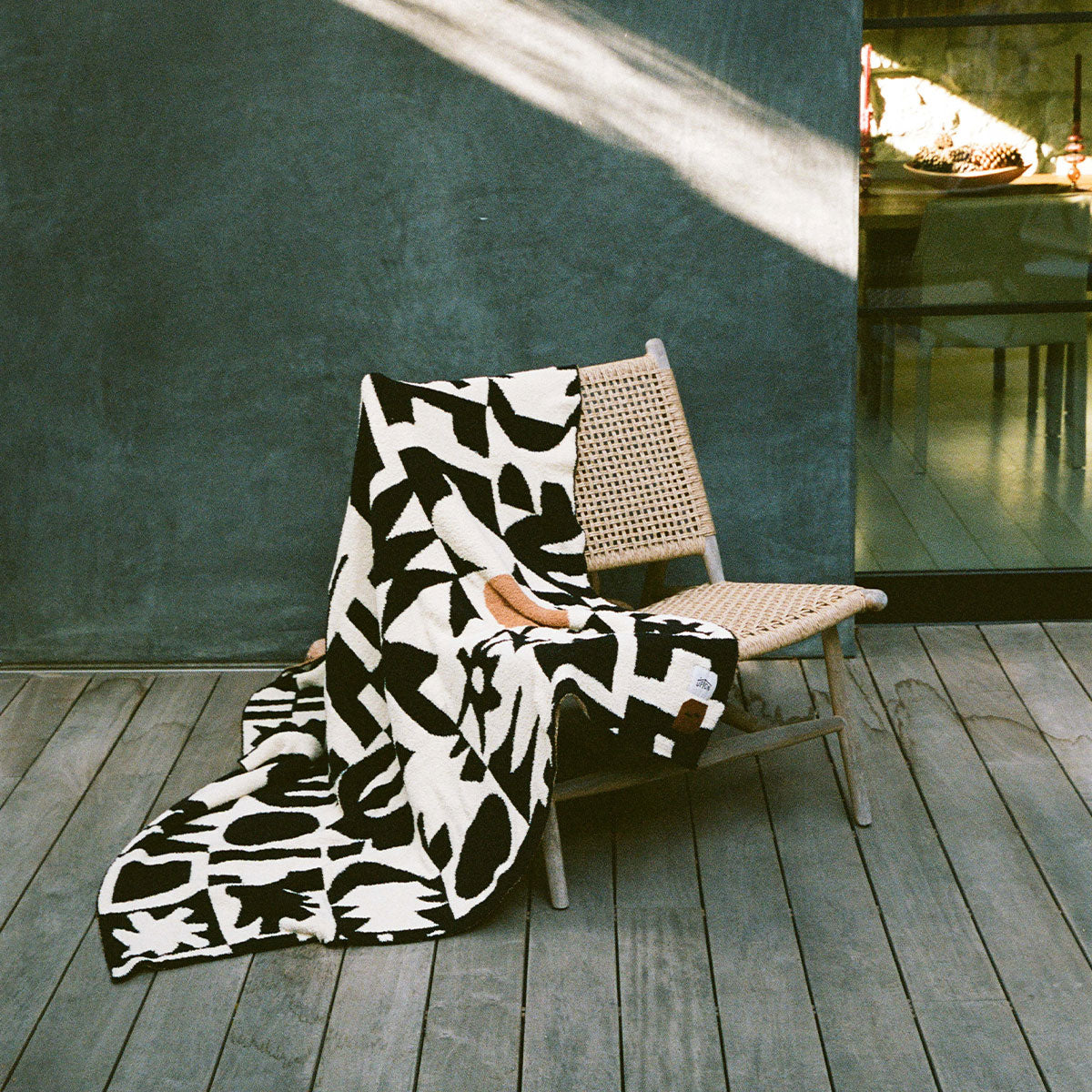A woven chair sits on a wooden deck with a black and white patterned blanket draped over it; sunlight and shadows fall across the scene near a glass door.