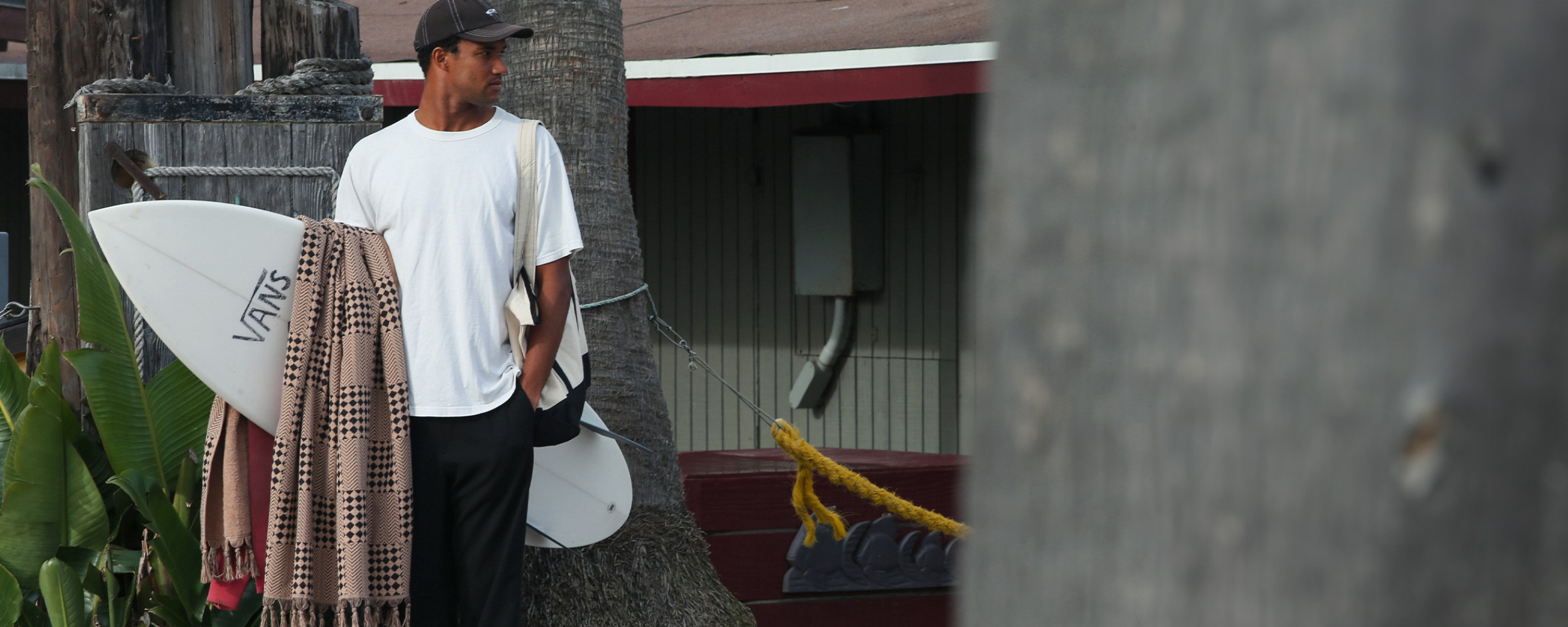 A man in a white t-shirt and cap stands outdoors holding a surfboard marked VANS and a striped towel, looking to his left, with trees and a building in the background.