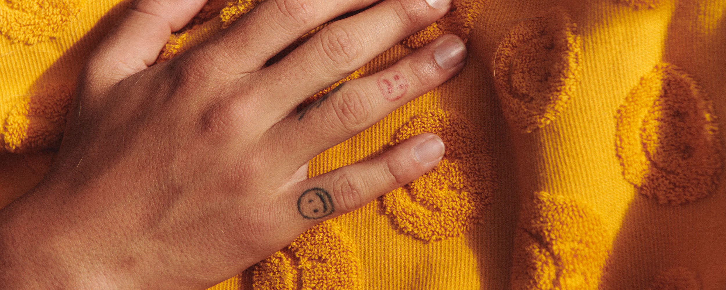 A hand with small smiley face tattoos on the fingers rests on a textured yellow fabric decorated with raised smiley face patterns.