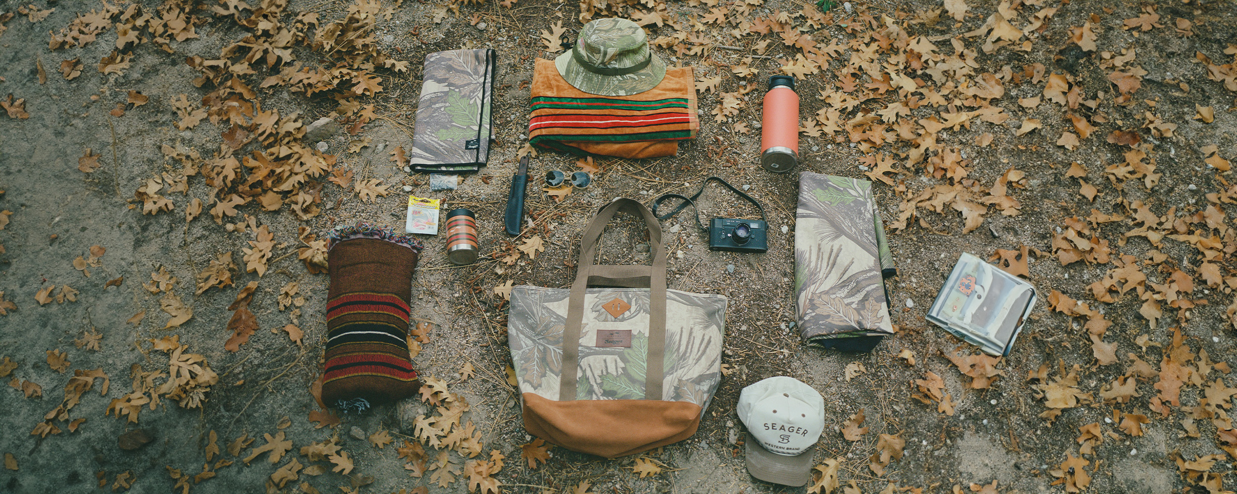 A flat lay of camping gear on autumn leaves, including a hat, tote bag, striped blanket, water bottle, camera, sunglasses, book, snacks, rope, and a Seager cap.