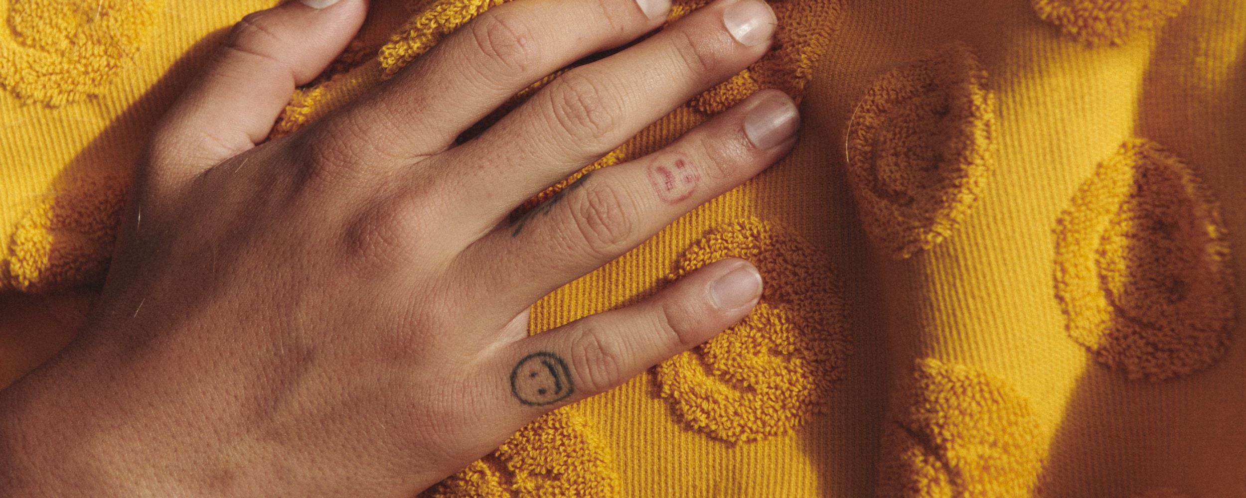 A hand rests on a yellow textured fabric with embroidered smiley faces. The fingers have small tattoos, including two smiley faces on the index and middle finger.