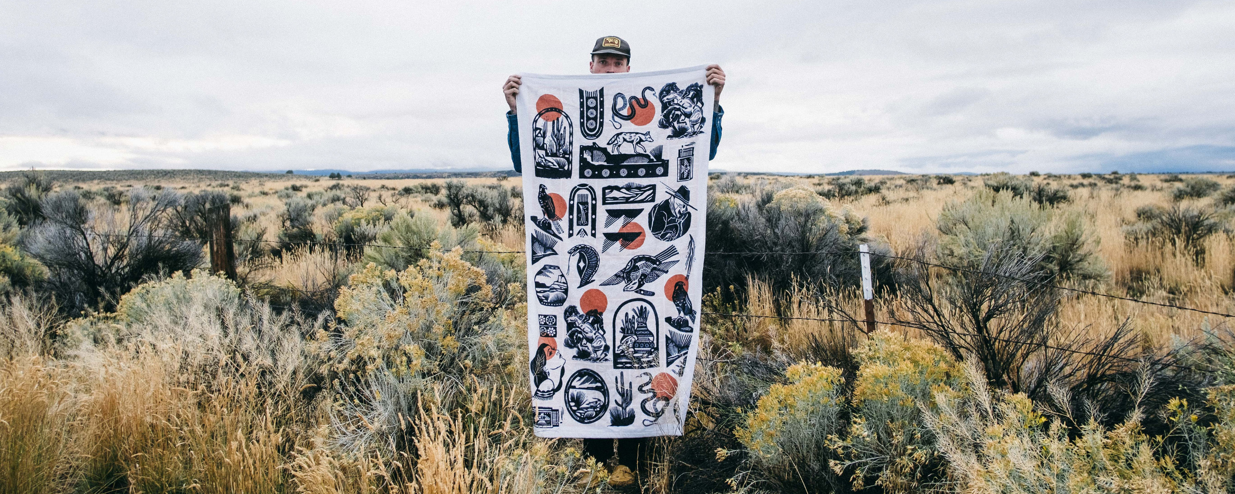 A person stands in a grassy, open field holding a large white cloth with black and red illustrations, partially hiding their face and body. The sky is overcast and the landscape is wild and natural.