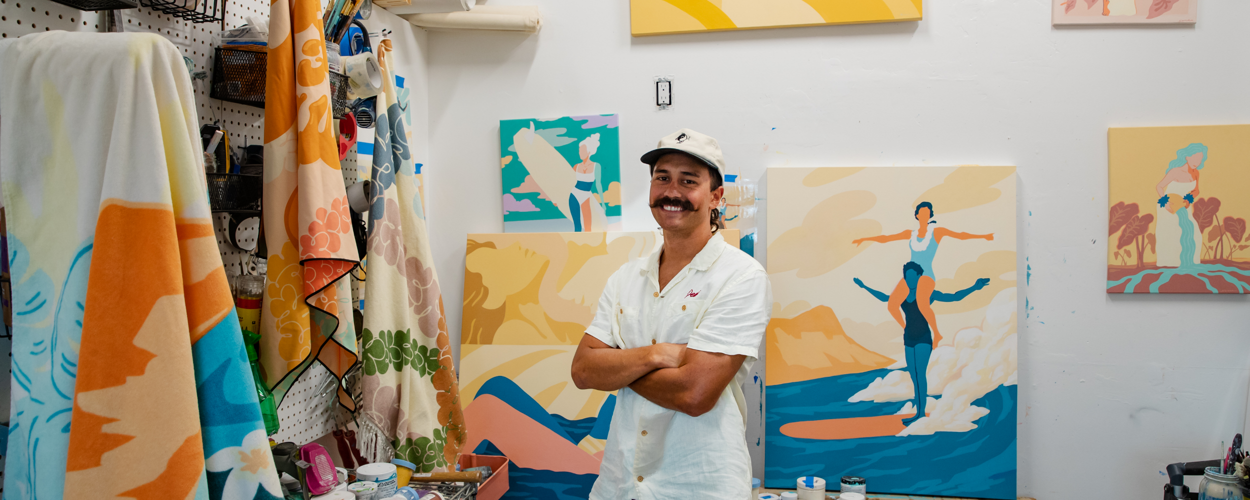 A man in a white shirt and cap stands smiling with arms crossed in an art studio, surrounded by colorful, abstract paintings and art supplies.