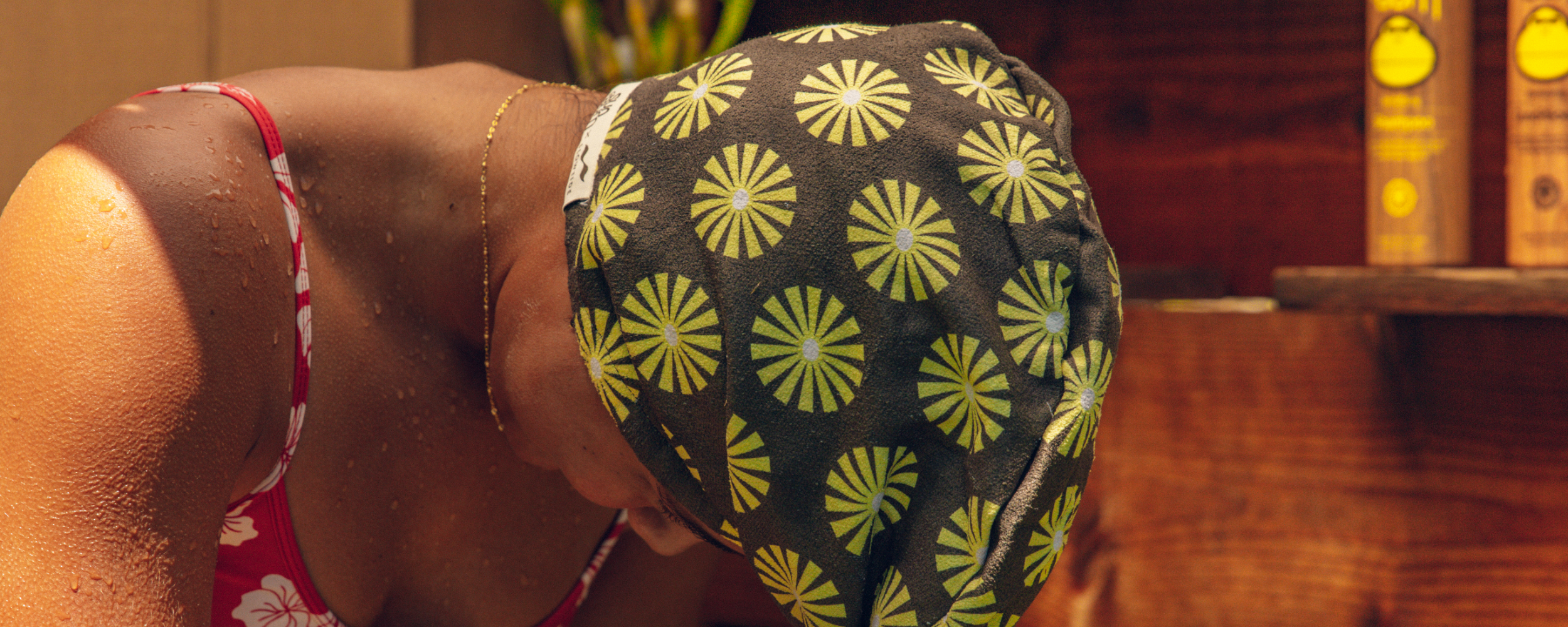 A person wearing a patterned black and green swim cap and a red swimsuit with white flowers leans forward, with water droplets on their skin and a wooden background behind them.