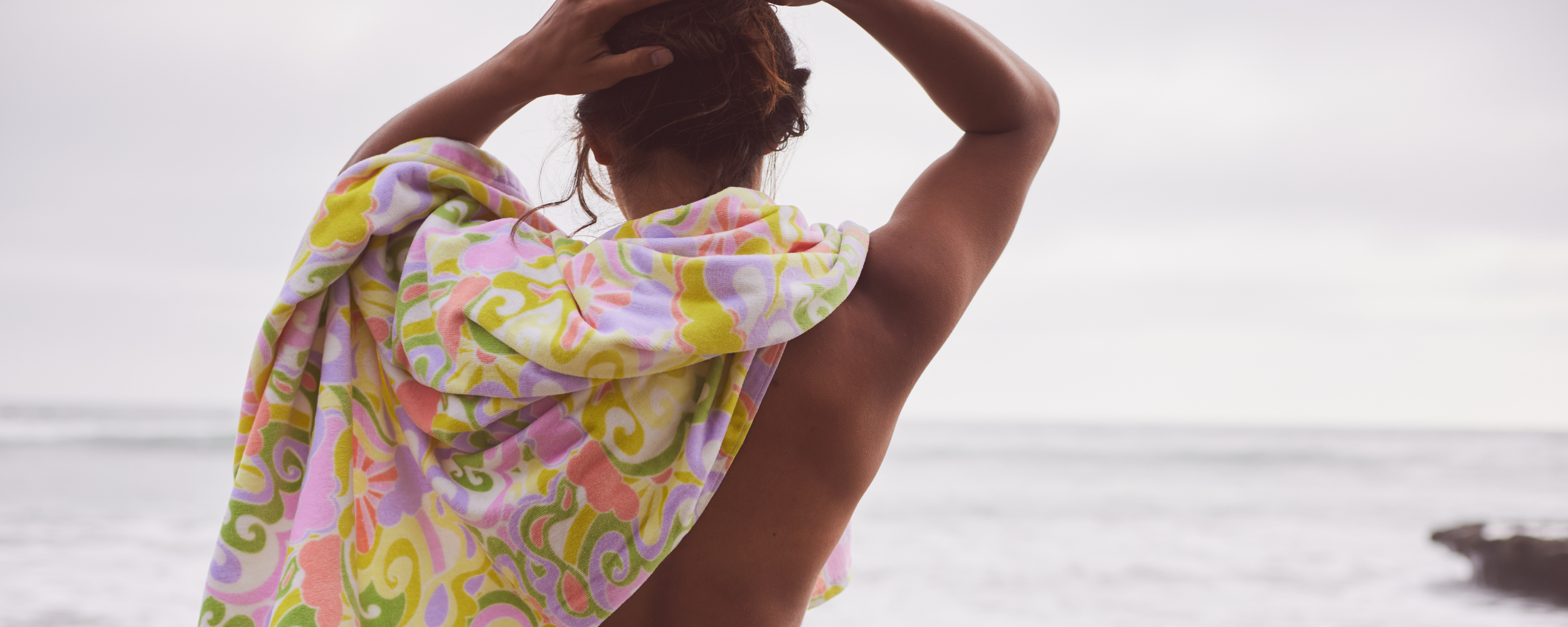A person with their back to the camera wraps a colorful towel around their shoulders while standing on a beach, facing the ocean on a cloudy day.