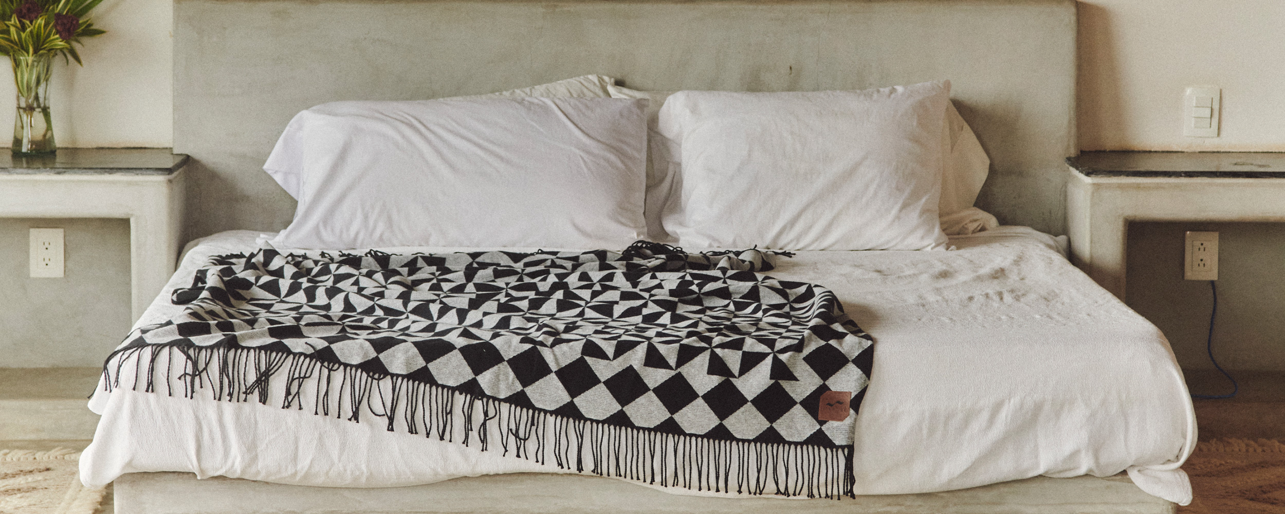 A neatly made bed with white pillows and a black-and-white geometric patterned throw blanket, in a minimalistic room with concrete nightstands and a vase of flowers.