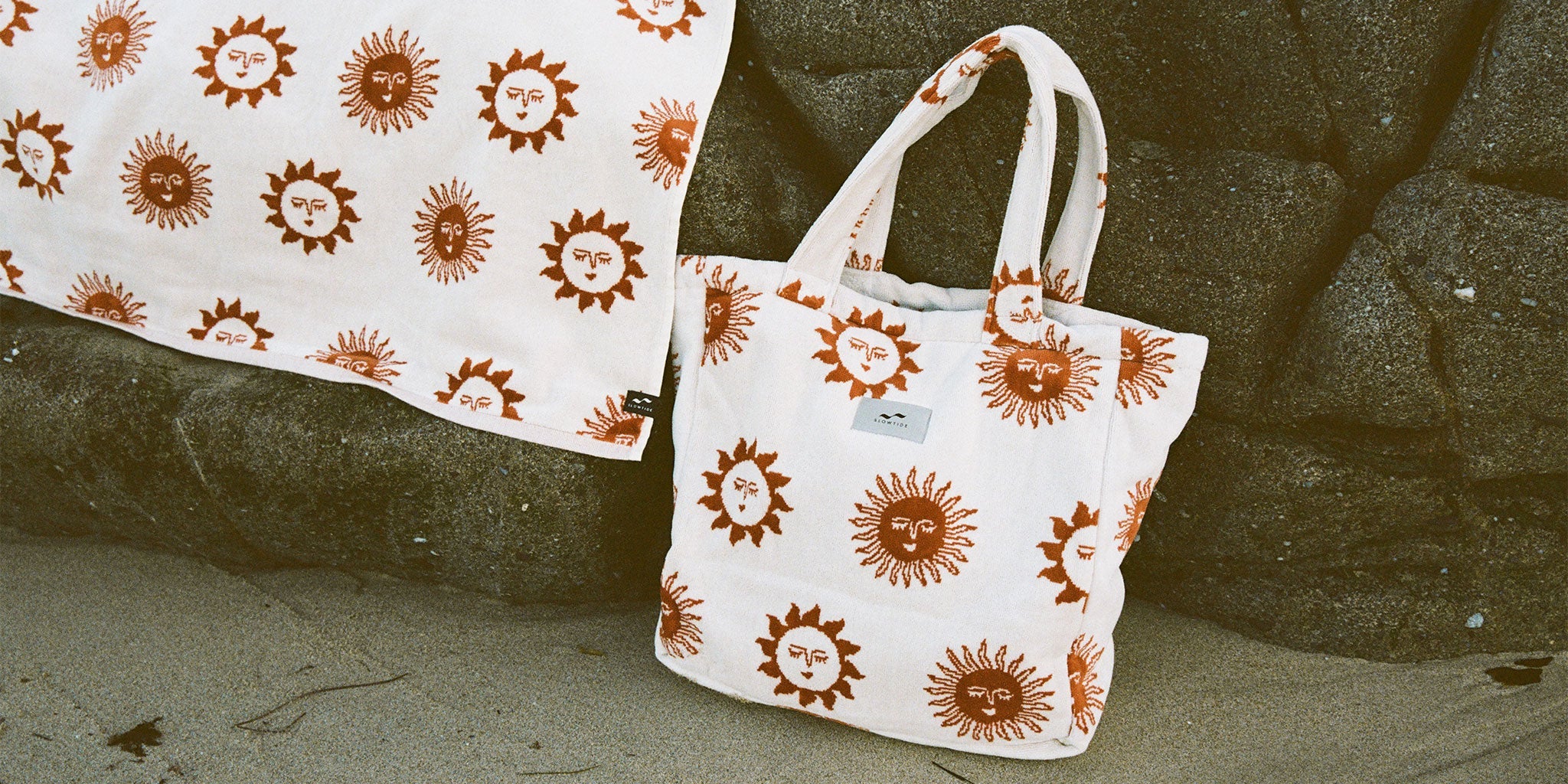 A white tote bag and cloth decorated with orange sun faces rest on sand and rocks, showcasing a repeating bold sunburst pattern with human-like expressions.