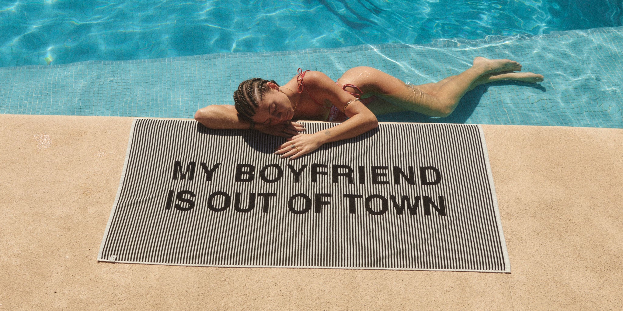 A woman in a bikini relaxes by a swimming pool, lying on a black-and-white striped towel that reads MY BOYFRIEND IS OUT OF TOWN in large black letters.