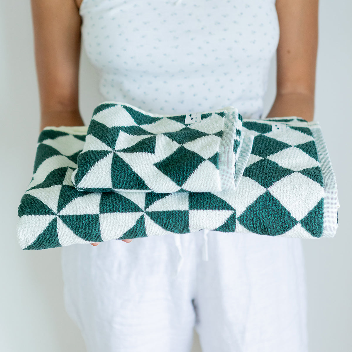 A person in a white sleeveless top and white pants holds a folded stack of green and white geometric-patterned towels against a plain background.