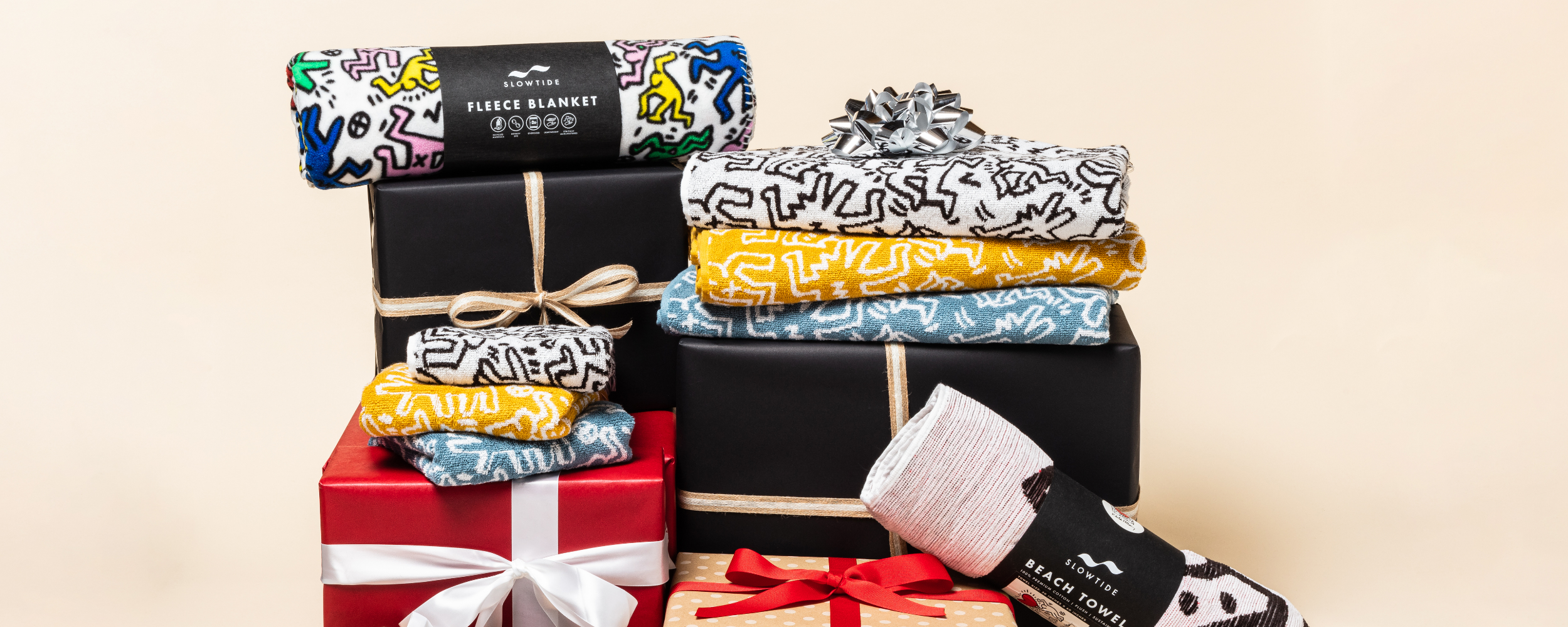 Colorful fleece blankets with abstract patterns are stacked on and next to wrapped gift boxes with ribbons and bows against a beige background. A pair of socks and smaller fabric items are also visible.