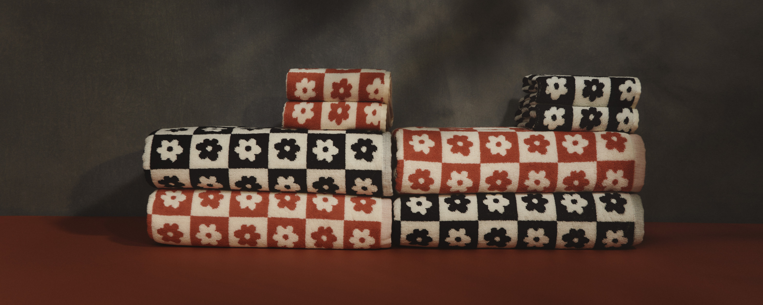 Stacked towels with checkered patterns in red and black, featuring white and black flower designs, arranged against a dark background on a red surface.