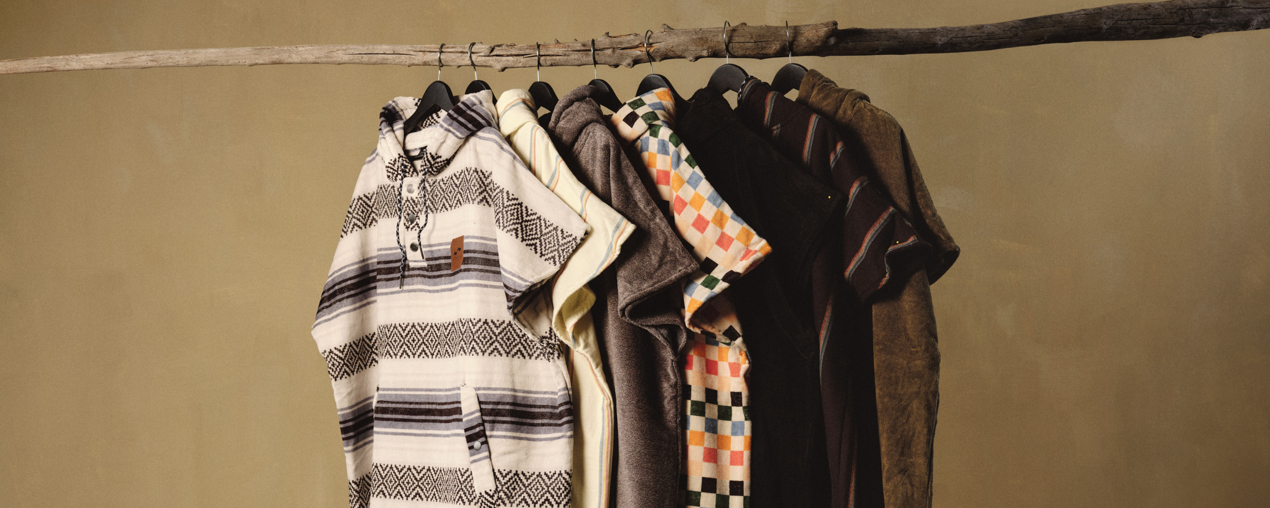 A row of various patterned and textured shirts on black hangers hangs from a rustic wooden branch against a plain beige background.
