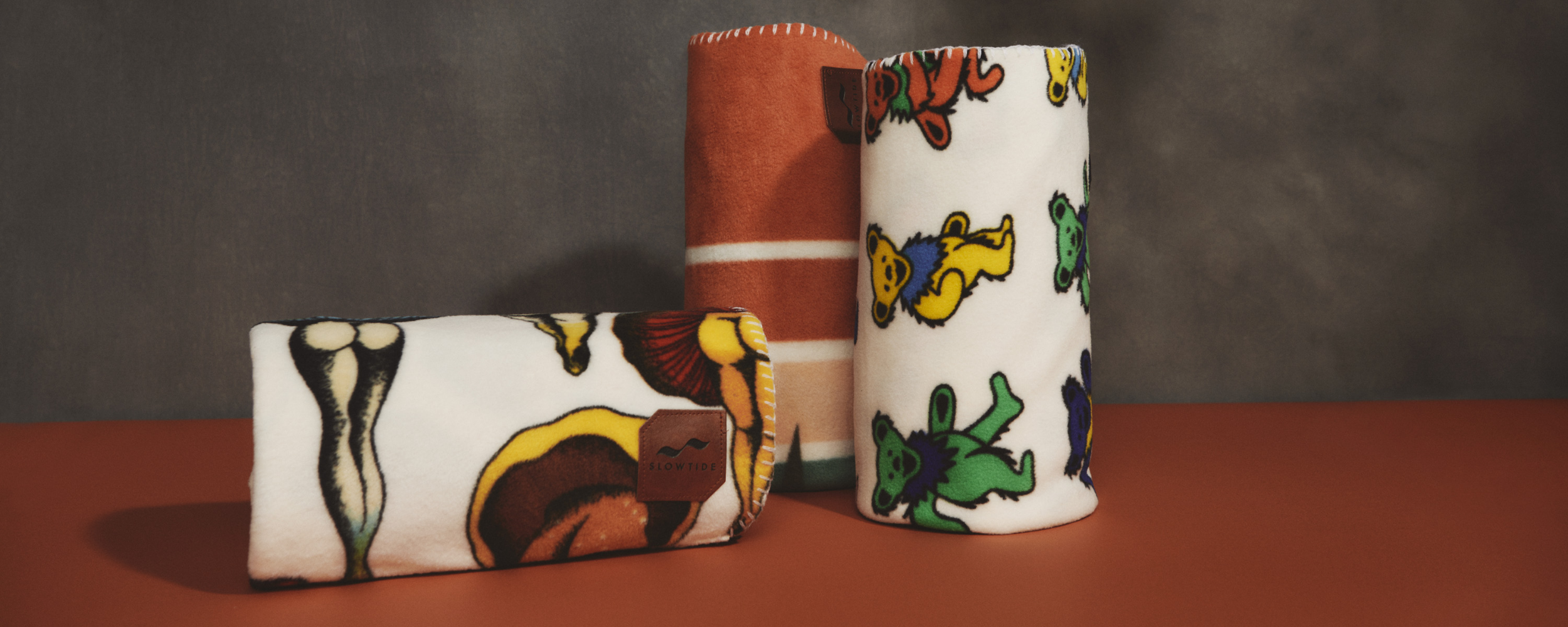 Three rolled fleece blankets on a brown surface; one with a skeleton and mushroom pattern, one with orange and white stripes, and one with colorful cartoon bears on a white background.