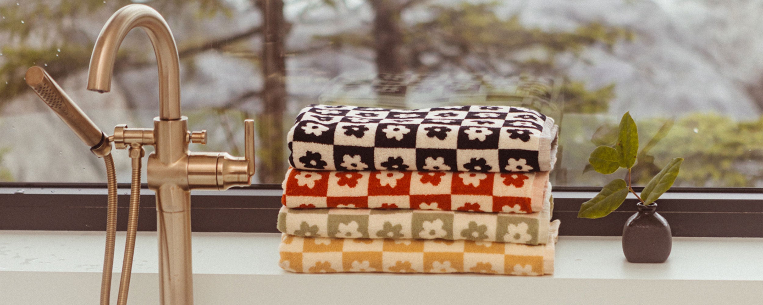 Four folded towels with a checkered and floral pattern in black, brown, yellow, and green sit on a windowsill next to a small potted plant and a gold faucet, with trees visible outside the window.