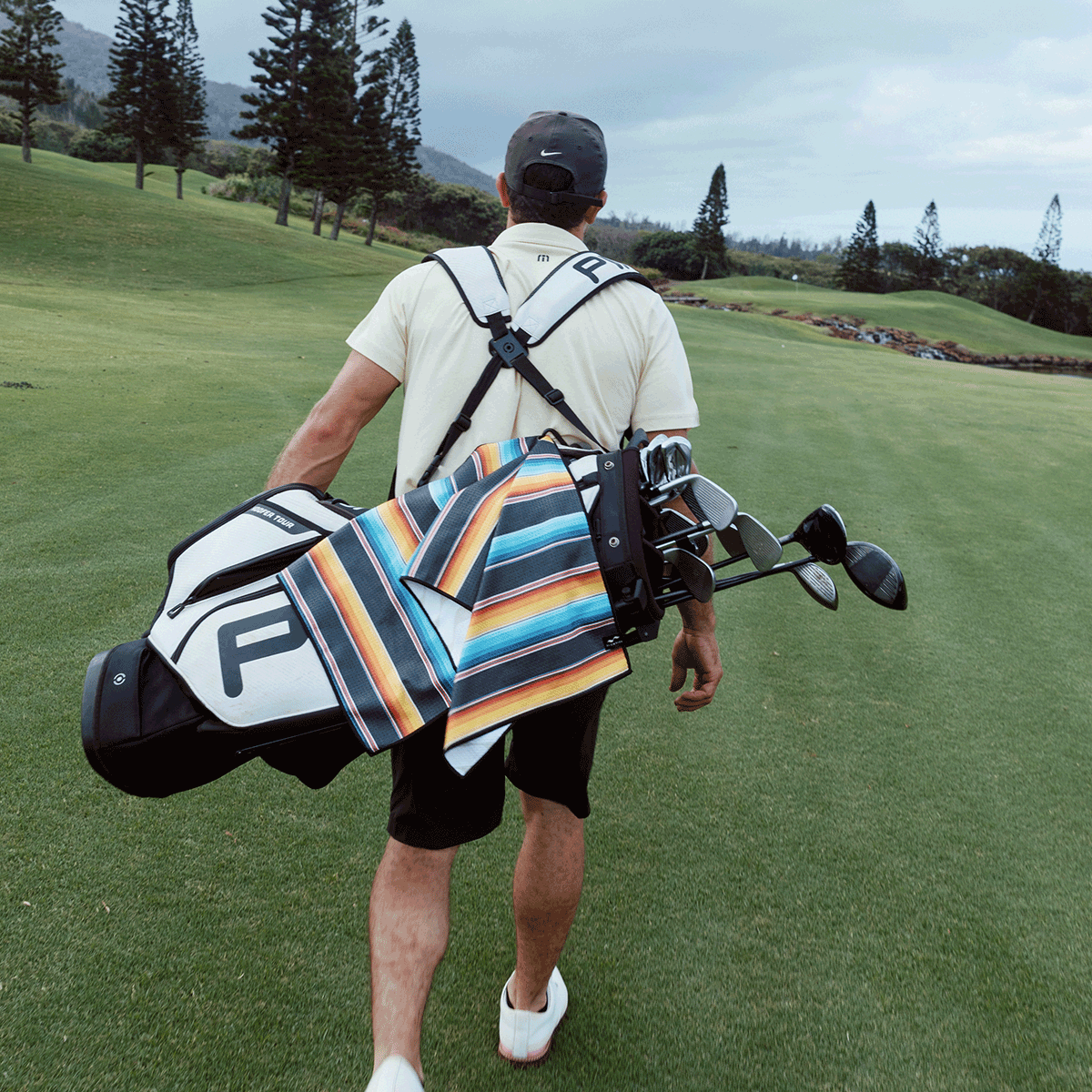 A man walks on a golf course carrying a golf bag with a colorful striped towel draped over it. Trees and hills are visible in the background under a cloudy sky.