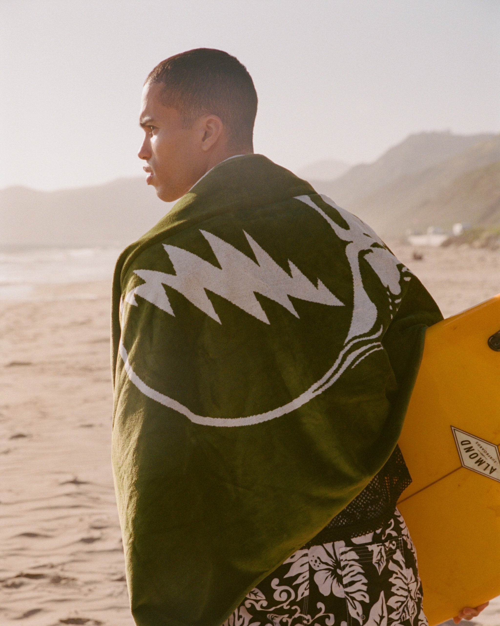 A person stands on a sandy beach, wrapped in a green towel with a white lightning bolt design, holding a yellow boogie board. Mountains and the ocean are visible in the background.