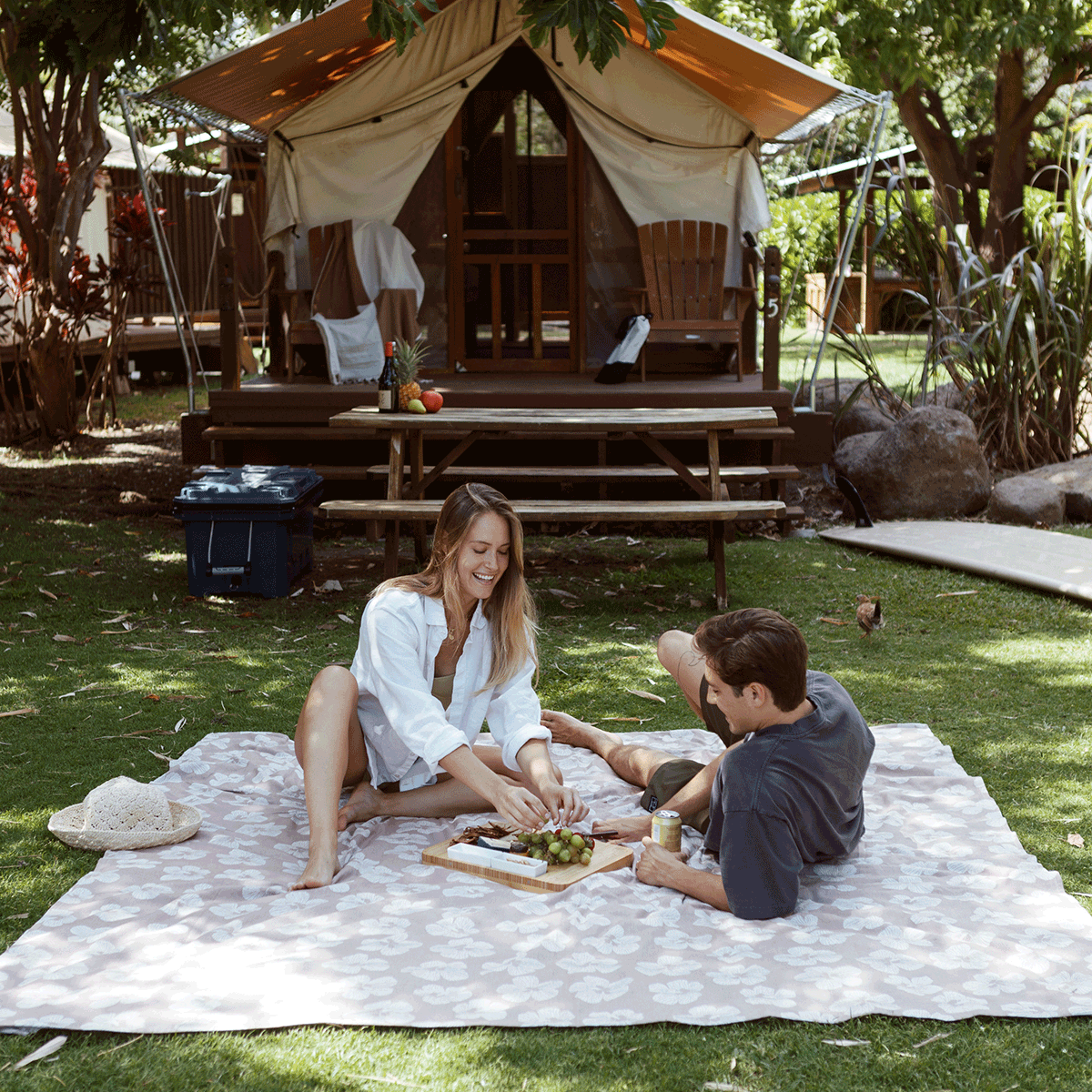 A woman and man enjoy a picnic on a blanket in front of a glamping tent, surrounded by trees and greenery on a sunny day. Food and drinks are laid out on a wooden tray between them.