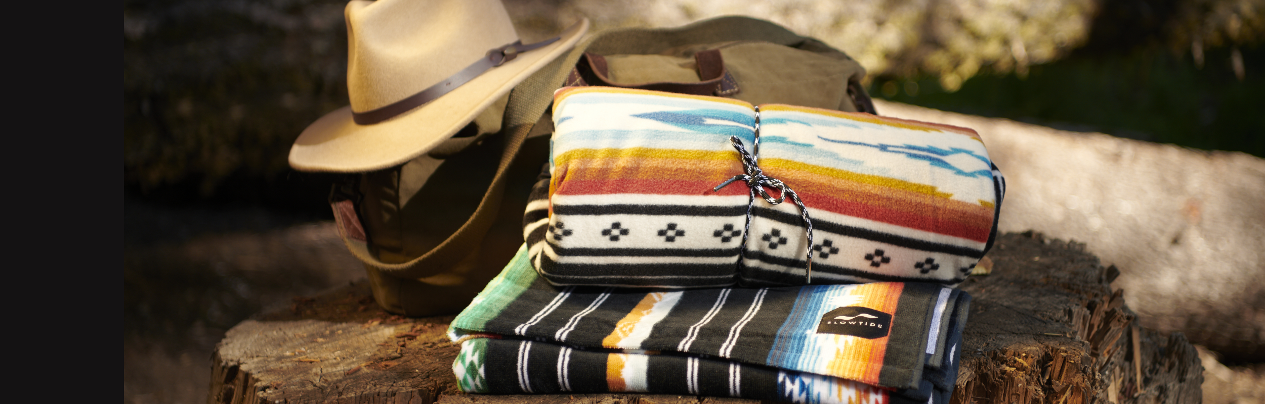A tan hat, a green bag, and two colorful patterned blankets are stacked on a tree stump outdoors, with sunlight highlighting the textures and colors.