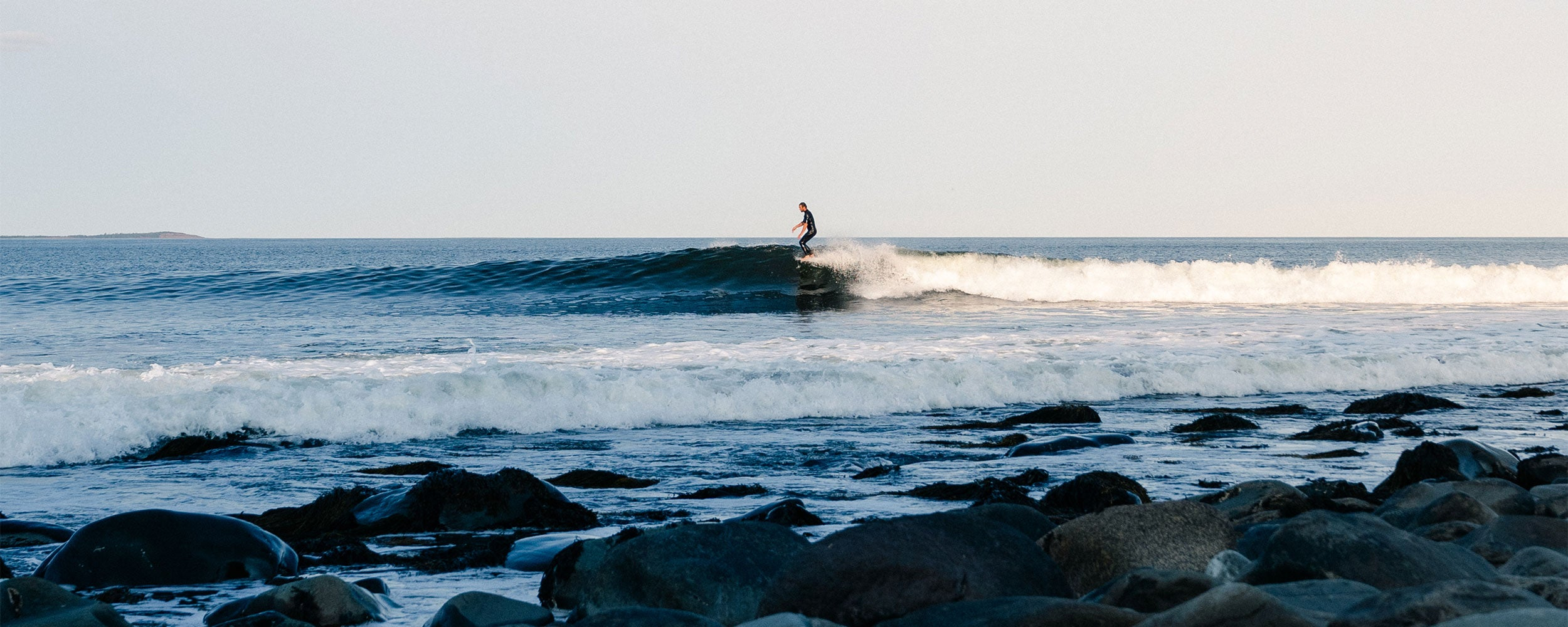 A person surfs on a gentle wave near the shore, with rocks and seaweed in the foreground and a calm, open ocean under a clear sky in the background.