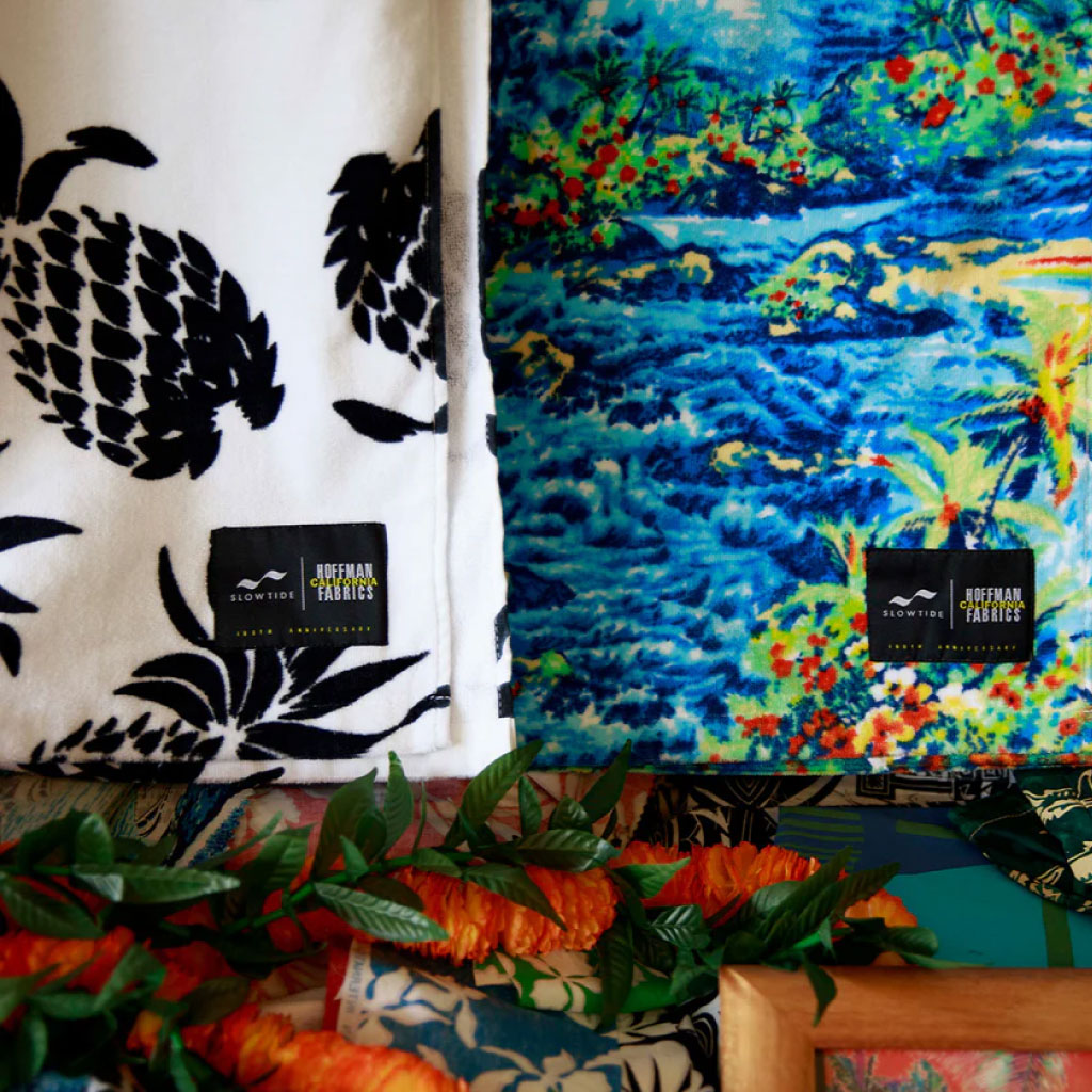 Two folded blankets: one with black pineapples on white, the other with a colorful tropical landscape. Both display fabric labels; artificial leaves and orange flowers are in the foreground.