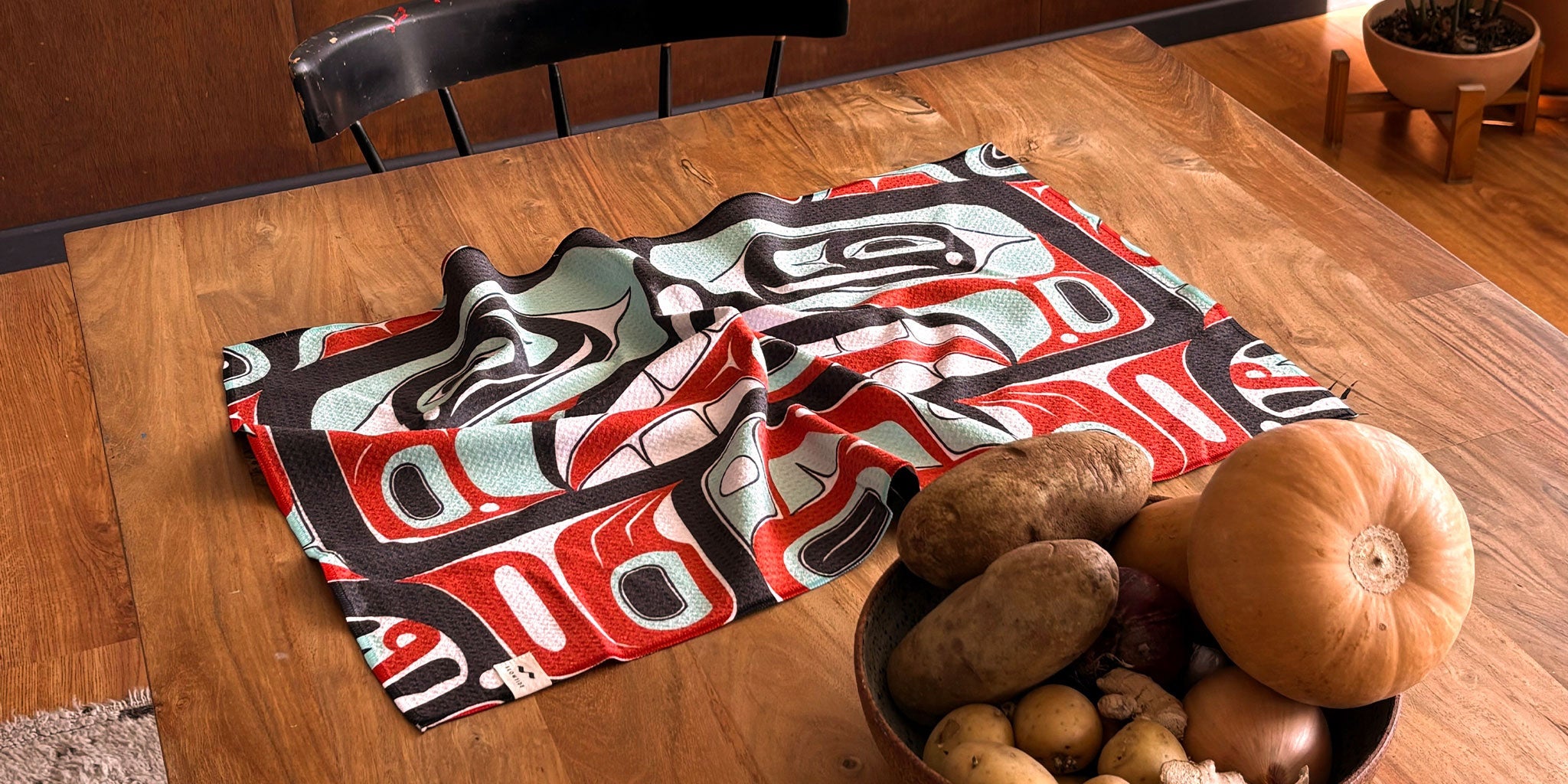 A wooden table with a colorful, Indigenous-style patterned cloth and a bowl filled with potatoes, onions, garlic, and a pumpkin. A black chair and potted plant are in the background.