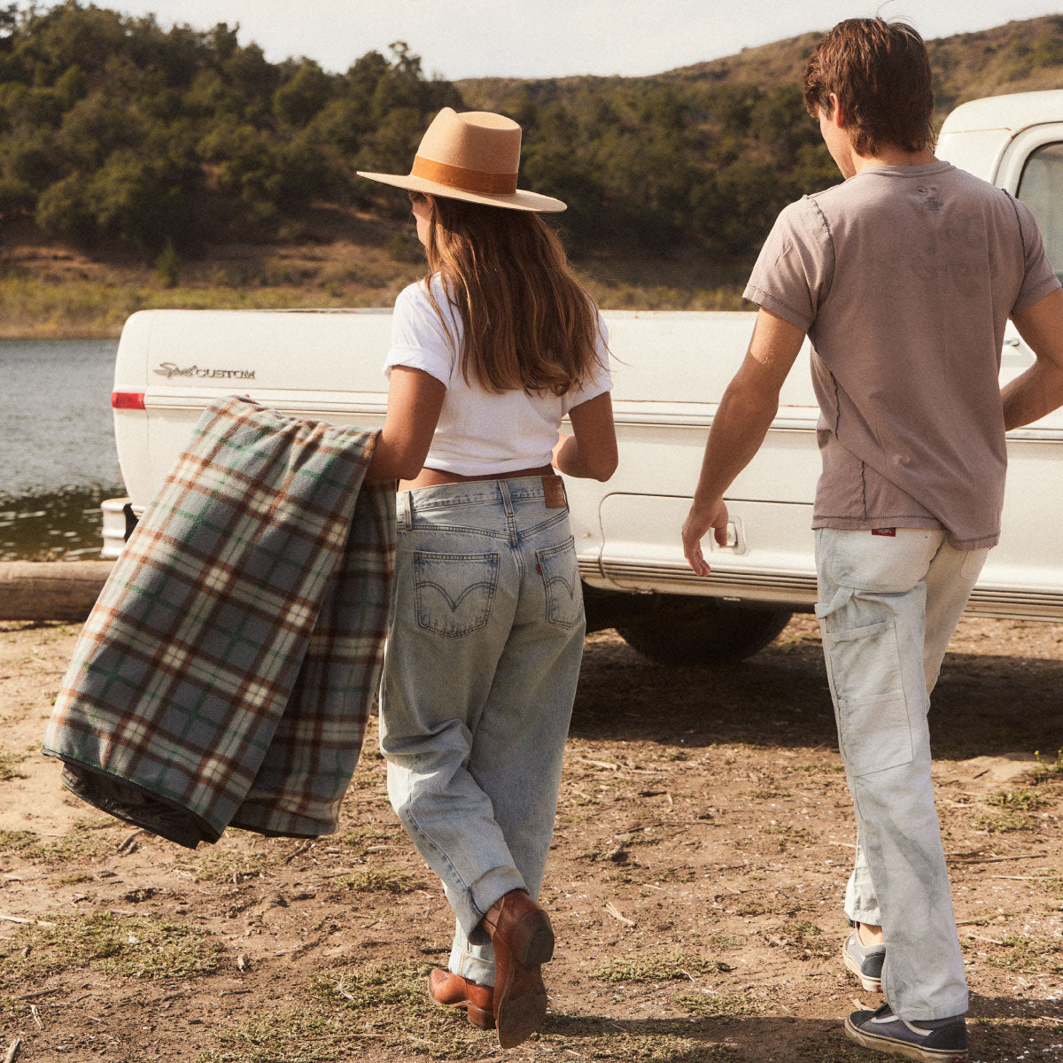 A woman in a hat and jeans carries a plaid blanket, walking with a man near a white pickup truck by a lake, with trees and hills in the background.