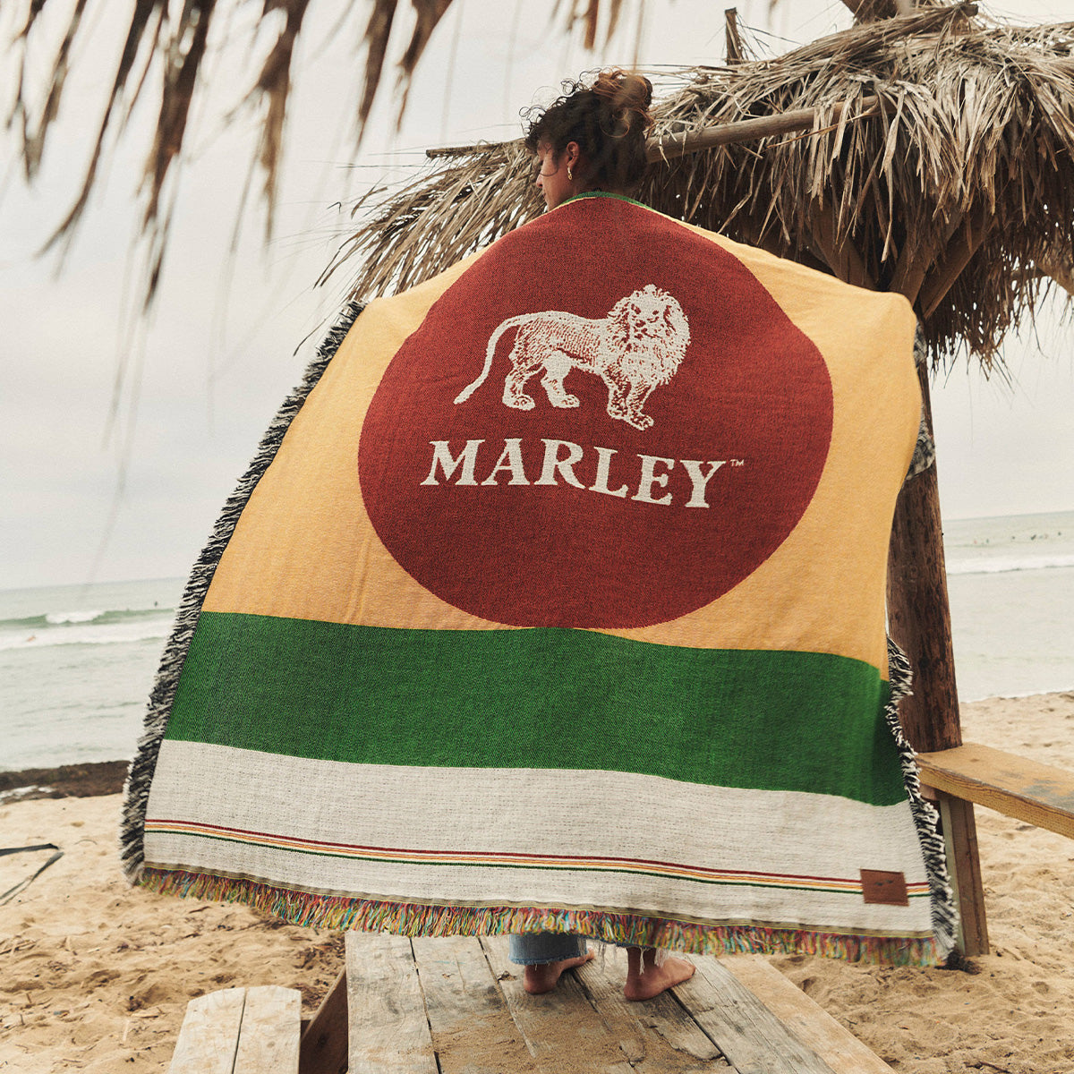 A person stands on a sandy beach, wrapped in a colorful blanket featuring a lion and the word MARLEY. Palm fronds and the ocean are visible in the background.