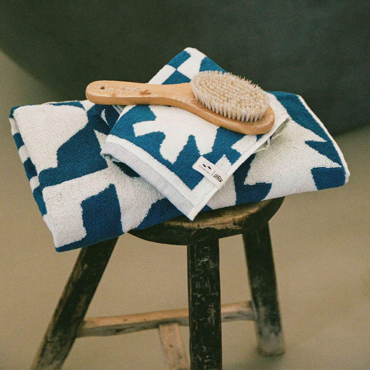 A wooden stool holds two blue and white patterned towels, neatly folded, with a wooden bath brush resting on top.