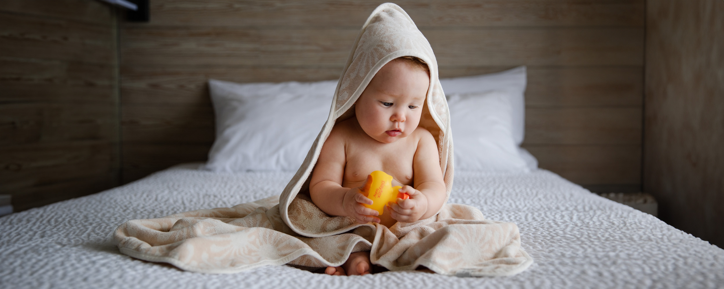 A baby wrapped in a beige towel with a hood sits on a bed, holding a yellow toy and looking down, with a pillow and wooden wall in the background.