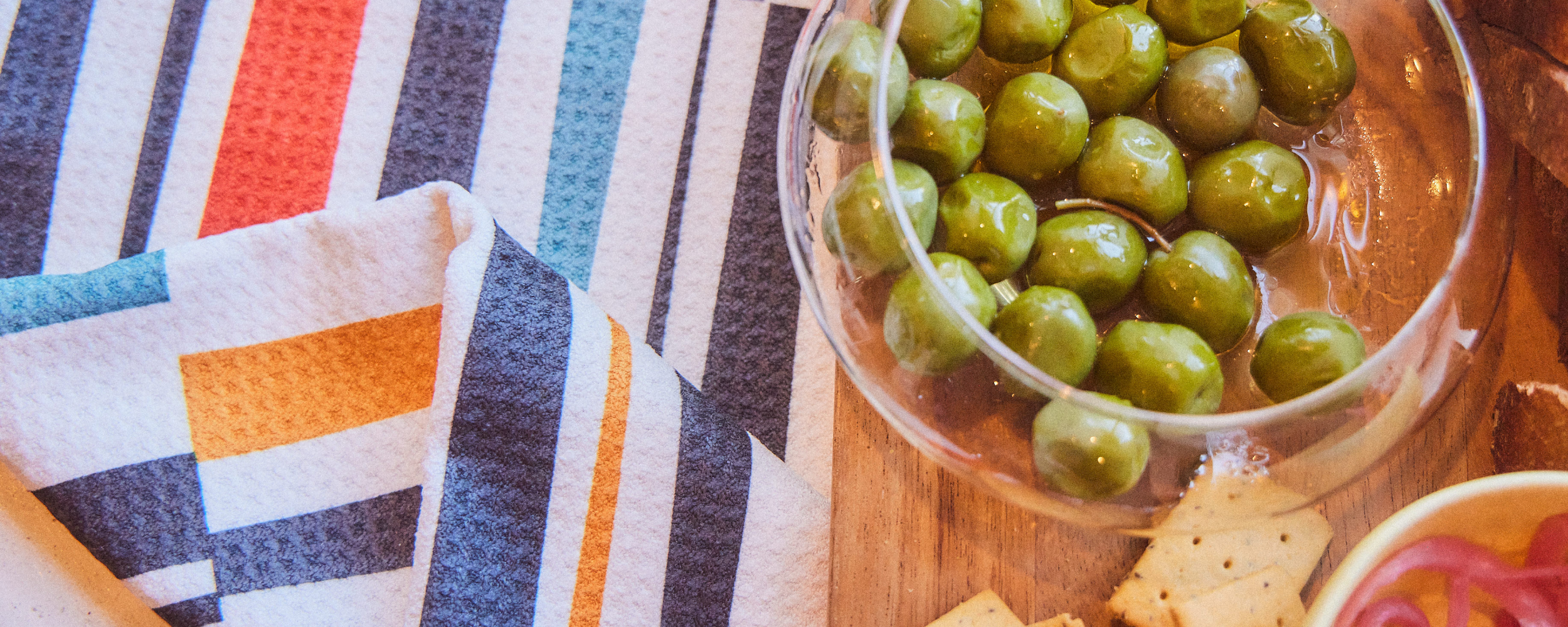A glass bowl of green olives sits on a wooden surface next to crackers and a colorful, striped napkin in shades of blue, orange, black, and white.