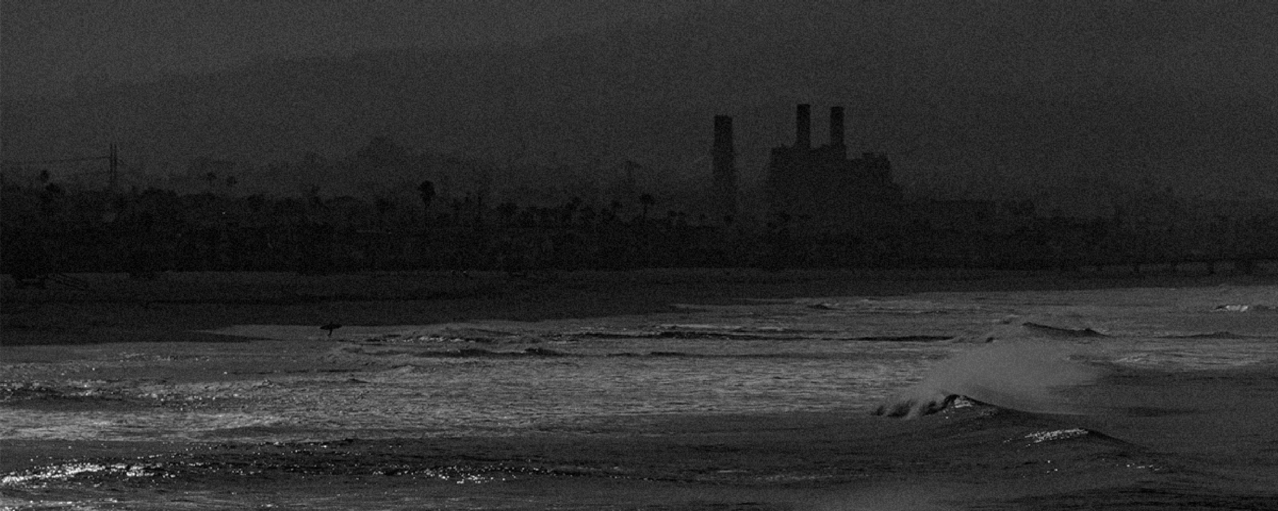 A dark, moody shoreline with waves crashing in the foreground and an industrial building with smokestacks silhouetted in the misty background.