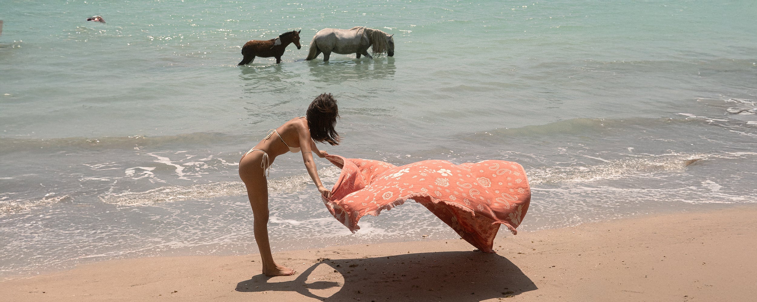 A woman in a bikini waves a patterned orange cloth on a sandy beach, while three horses stand in the shallow turquoise water behind her.